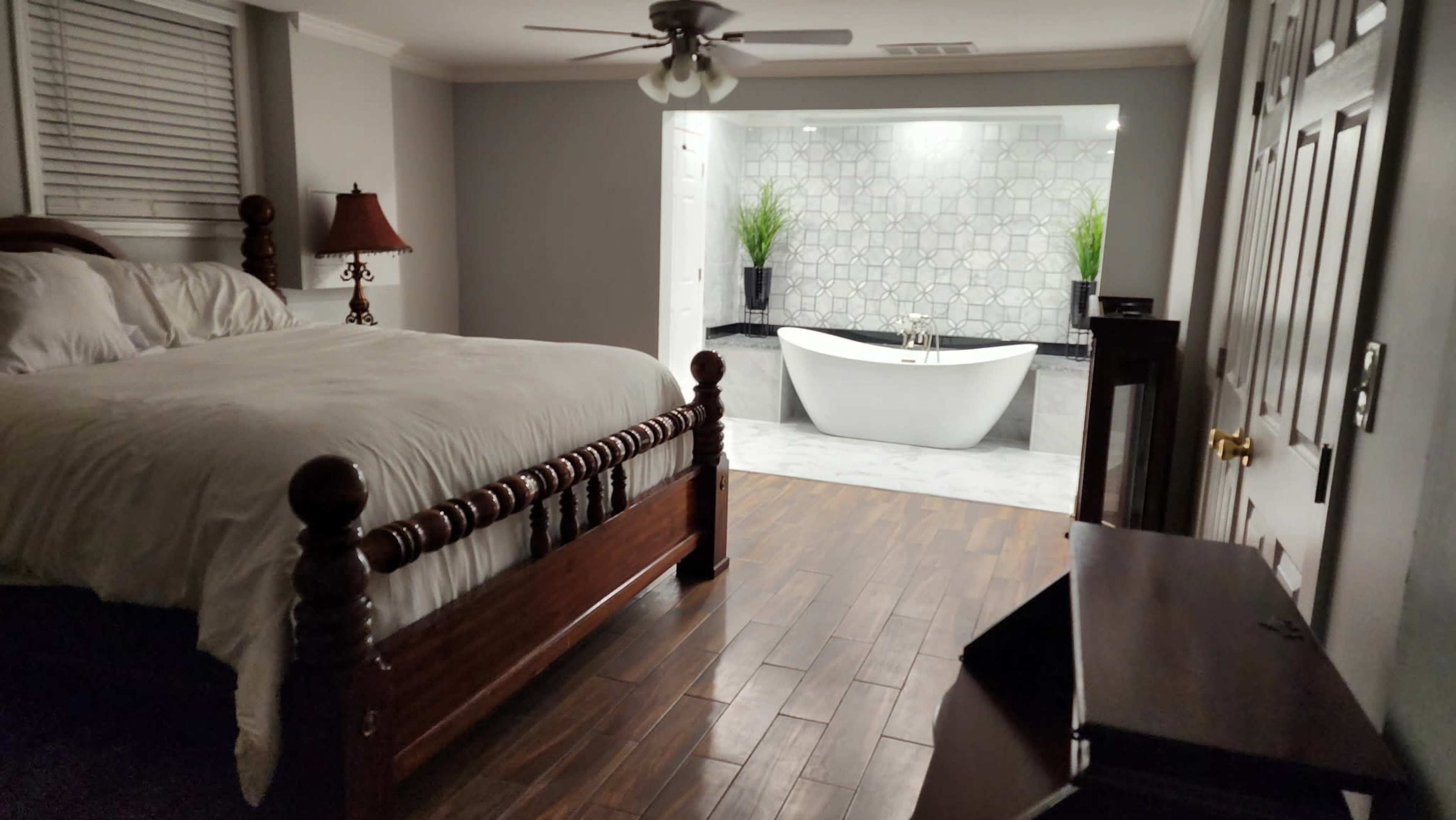 The image depicts a bedroom featuring a wooden bed with a white comforter, a lamp on a nightstand, and a modern bathroom area in the background with a freestanding bathtub and decorative plants.