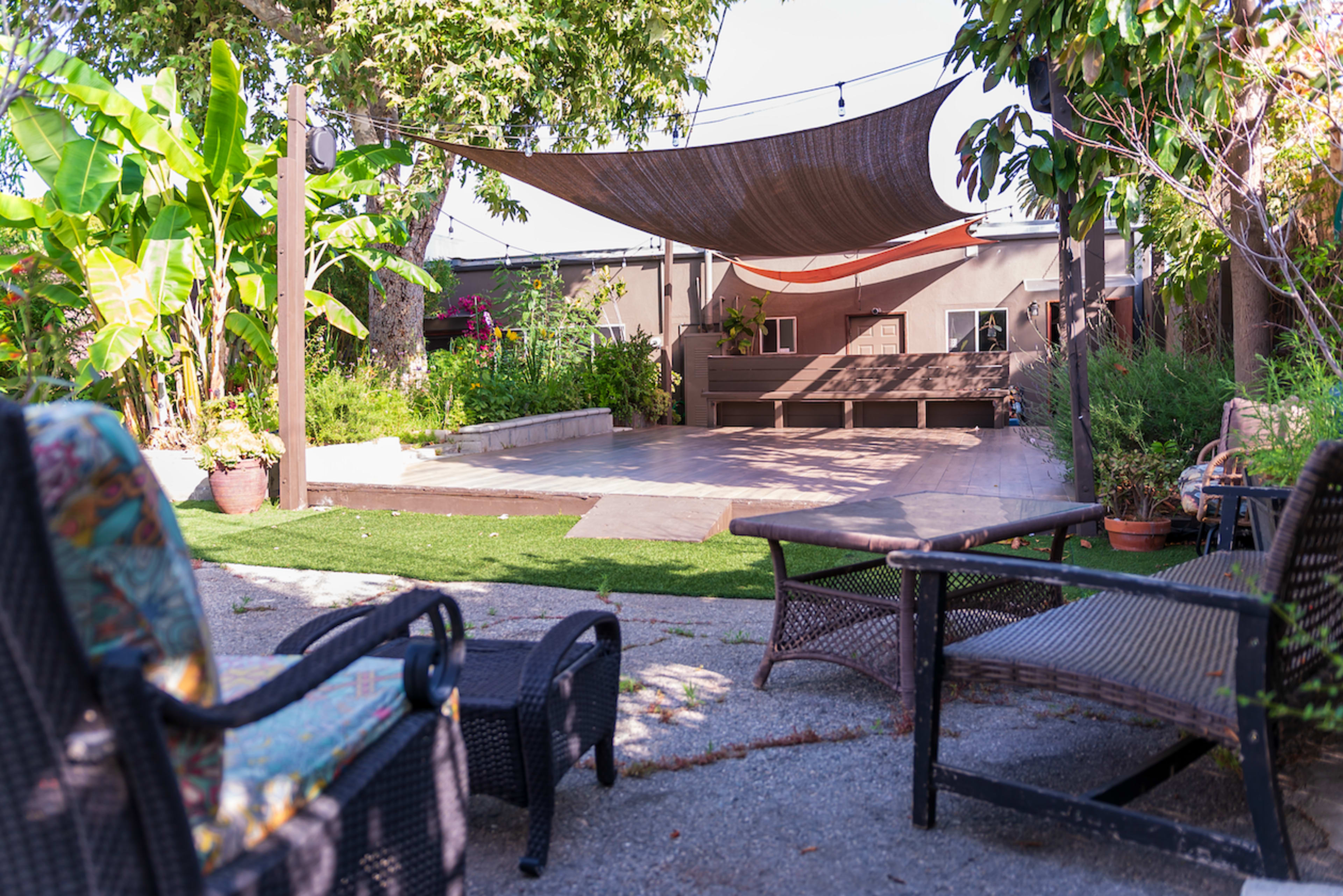 A backyard seating area features wicker chairs overlooking a paved space with a shade structure and surrounded by plants.