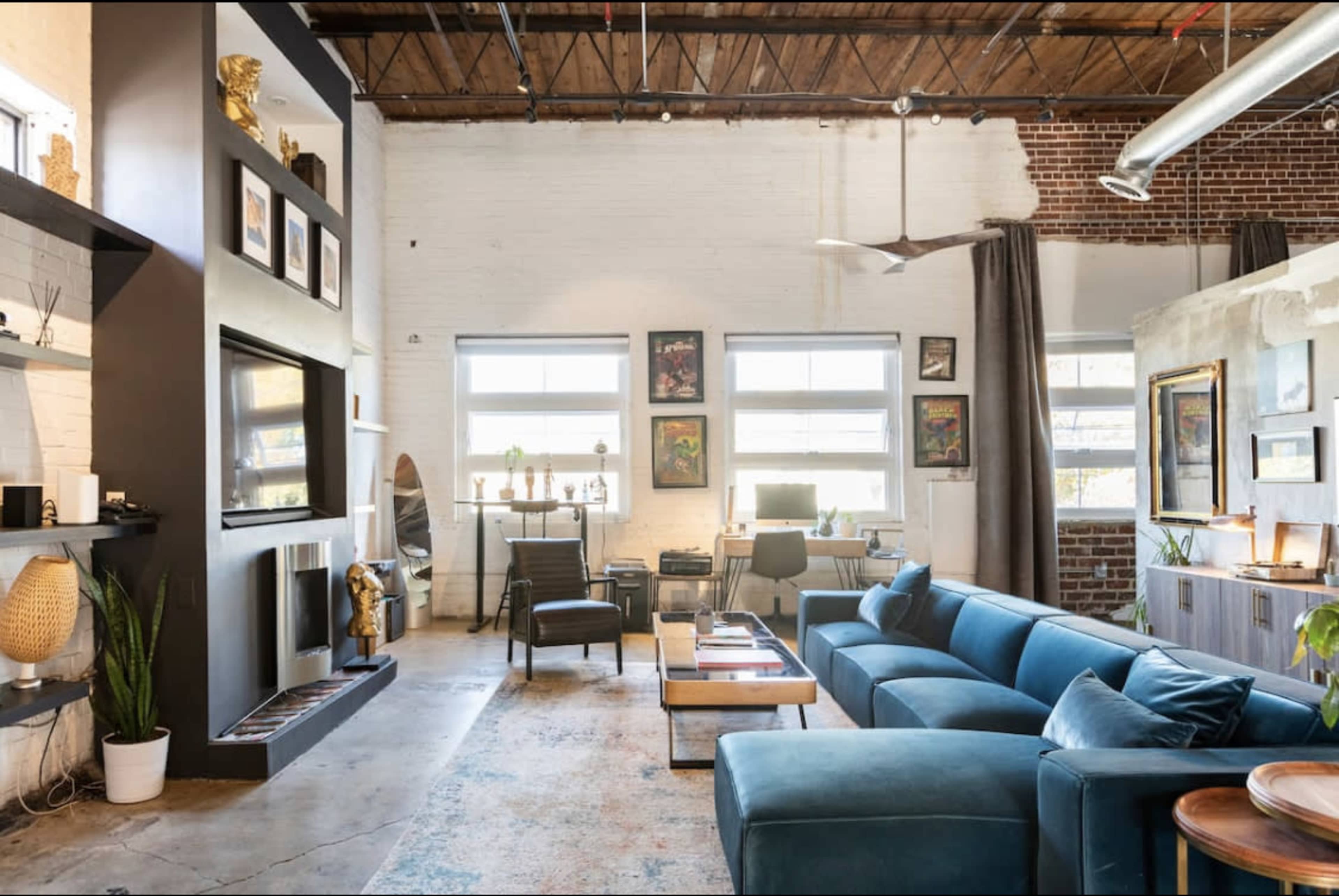 A spacious living area with a blue sectional sofa, large windows, and a mix of modern and vintage decor, including artwork and plants.