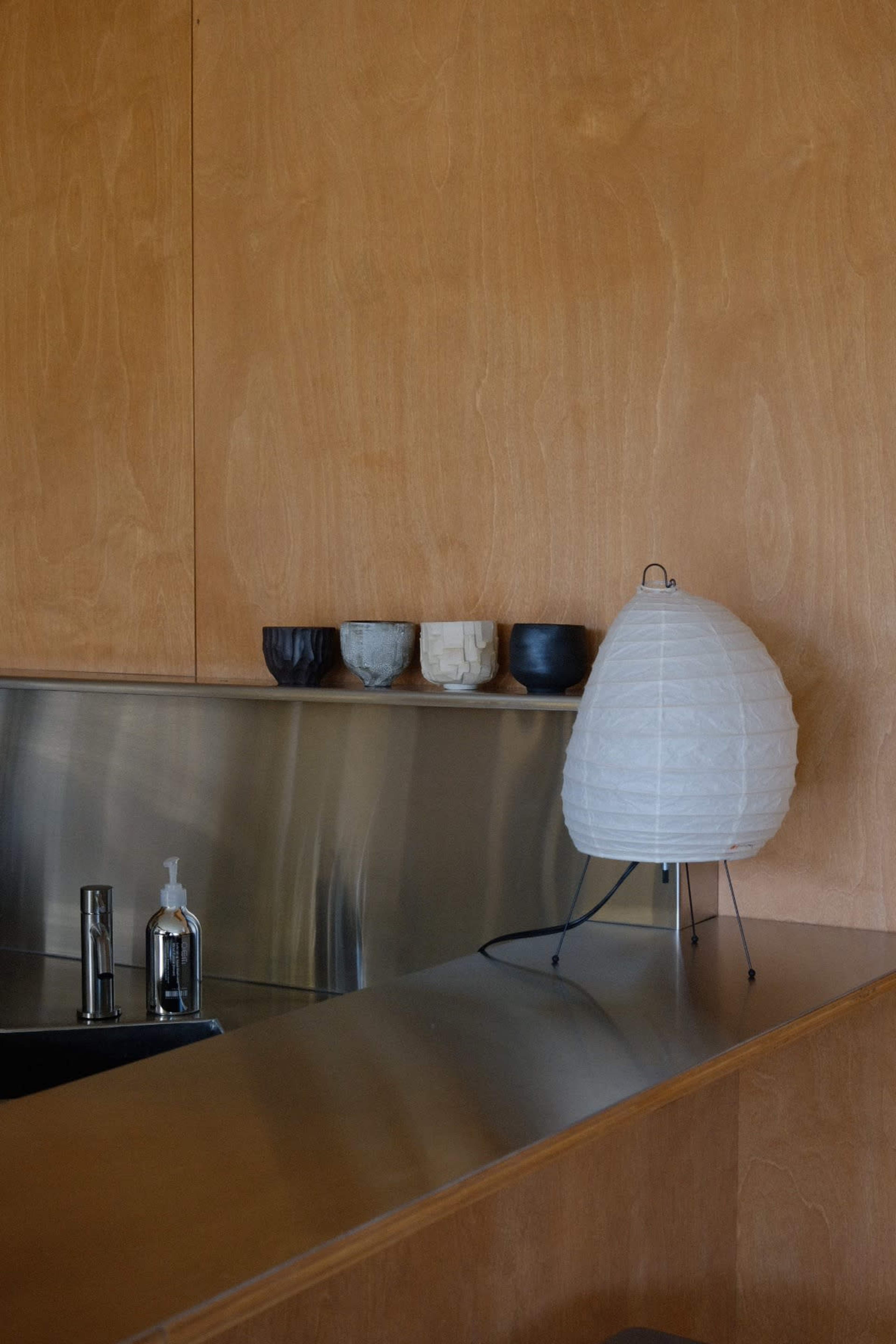 A minimalist kitchen counter features a metal surface, a soap dispenser, and a white lamp beside a row of small ceramic cups against a wooden wall.