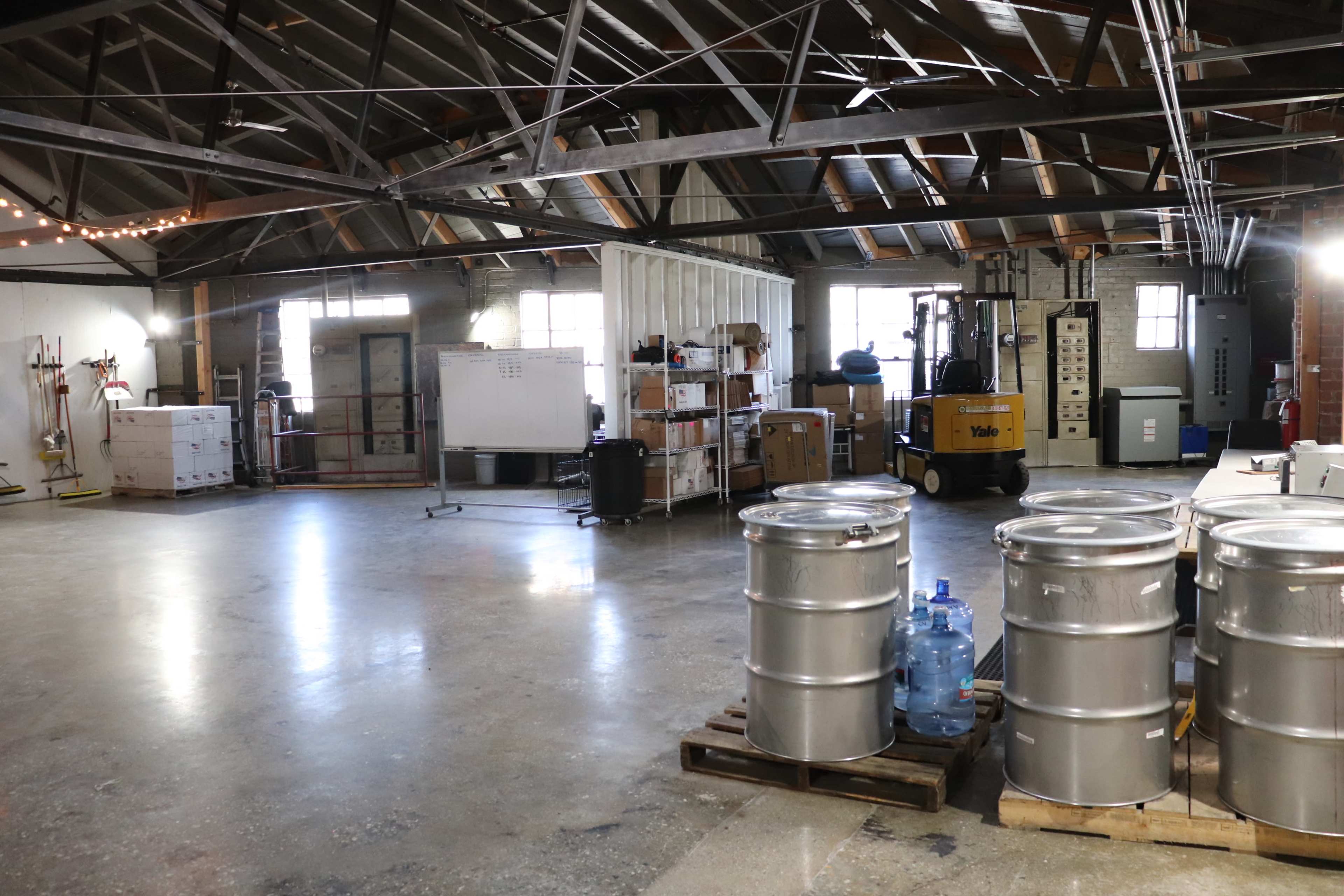 A spacious, industrial-style warehouse with large metal barrels, storage shelves, and a forklift in the background.
