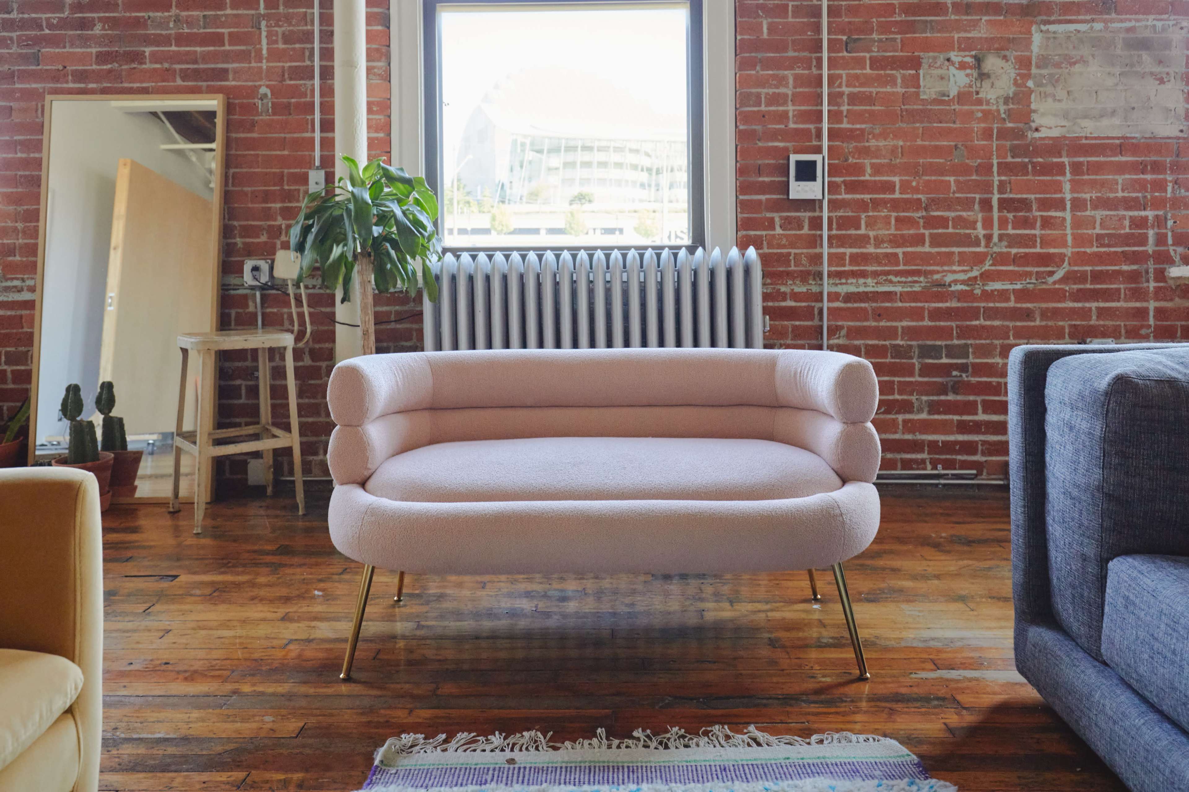 A pink sofa with gold legs sits on a wooden floor in a bright room featuring brick walls and a large window.