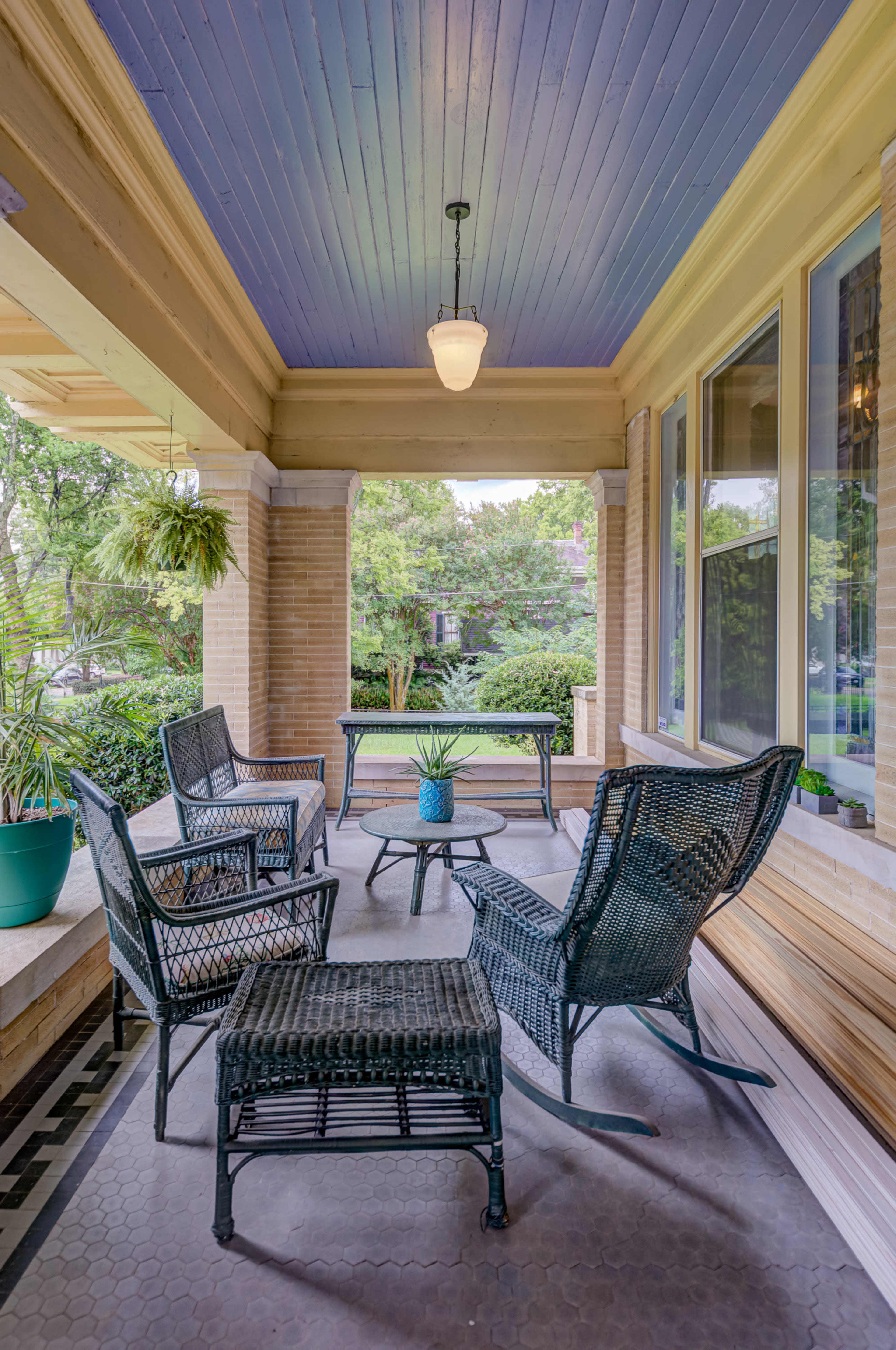 A covered porch features four wicker chairs arranged around a glass table with a blue plant pot in the center.