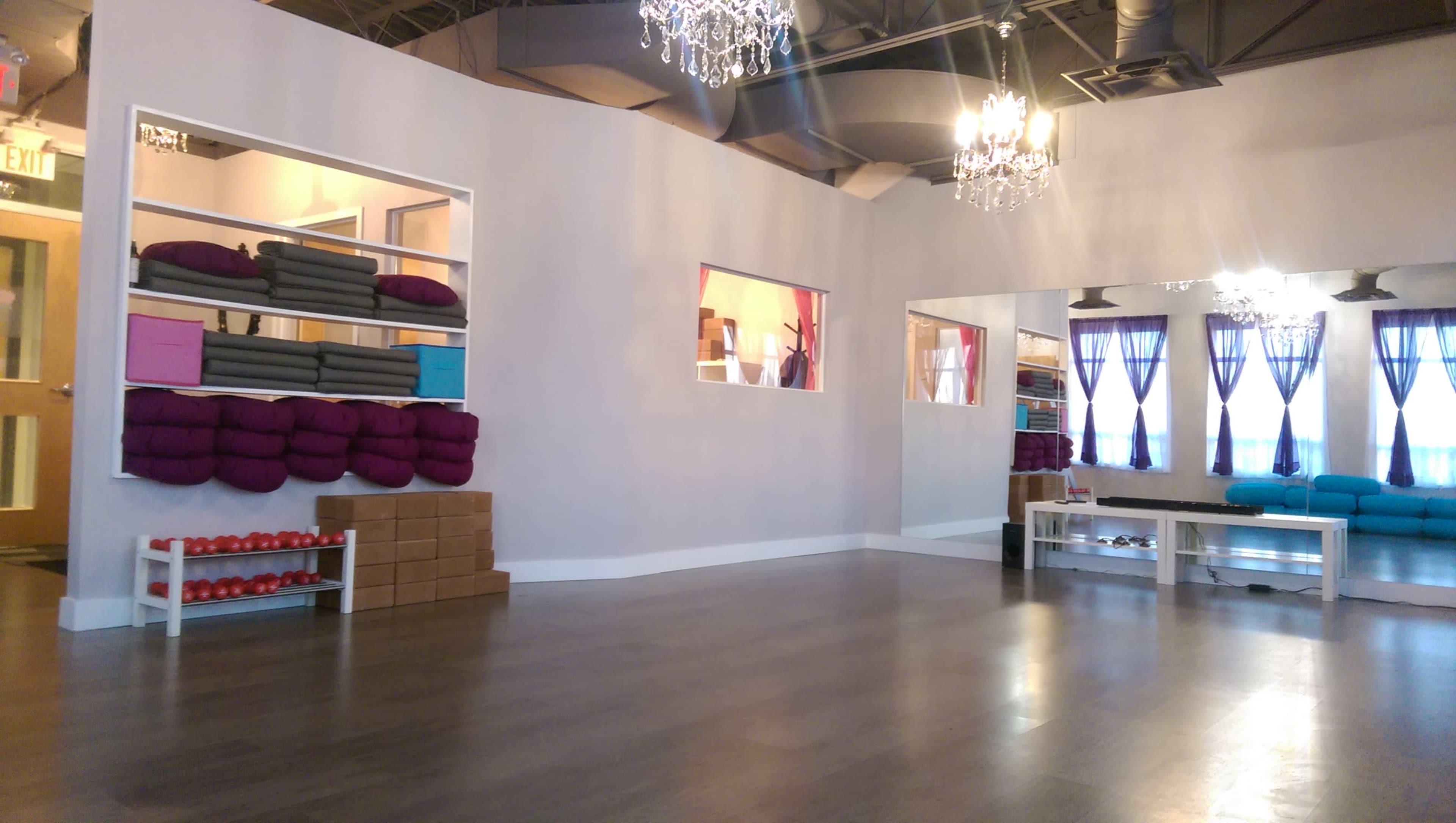 The interior of a yoga studio features large windows, hardwood floors, and neatly arranged exercise props along the walls.