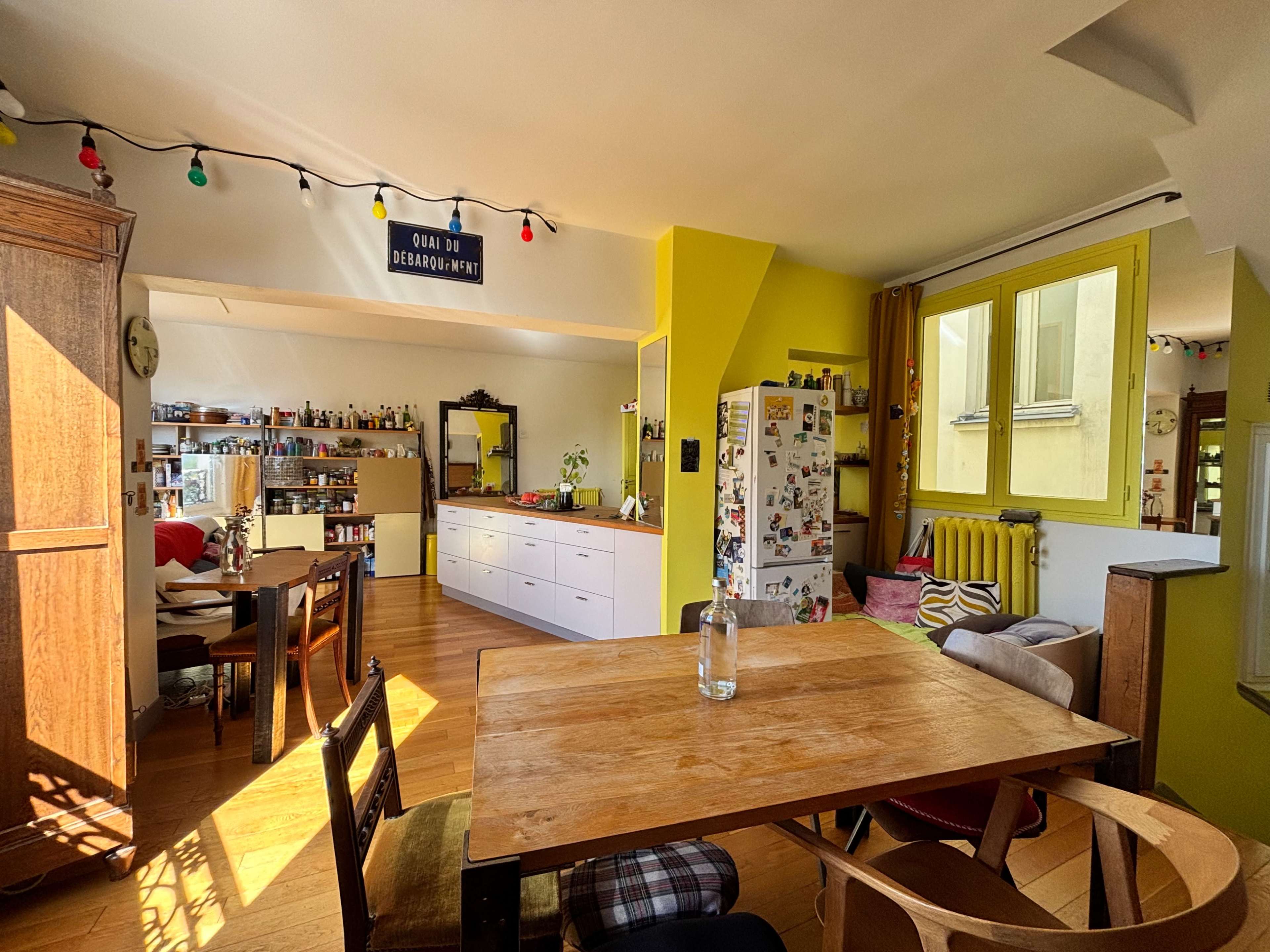 The image shows a bright, open kitchen and dining area with a wooden table, colorful walls, and various kitchen items and decorations.