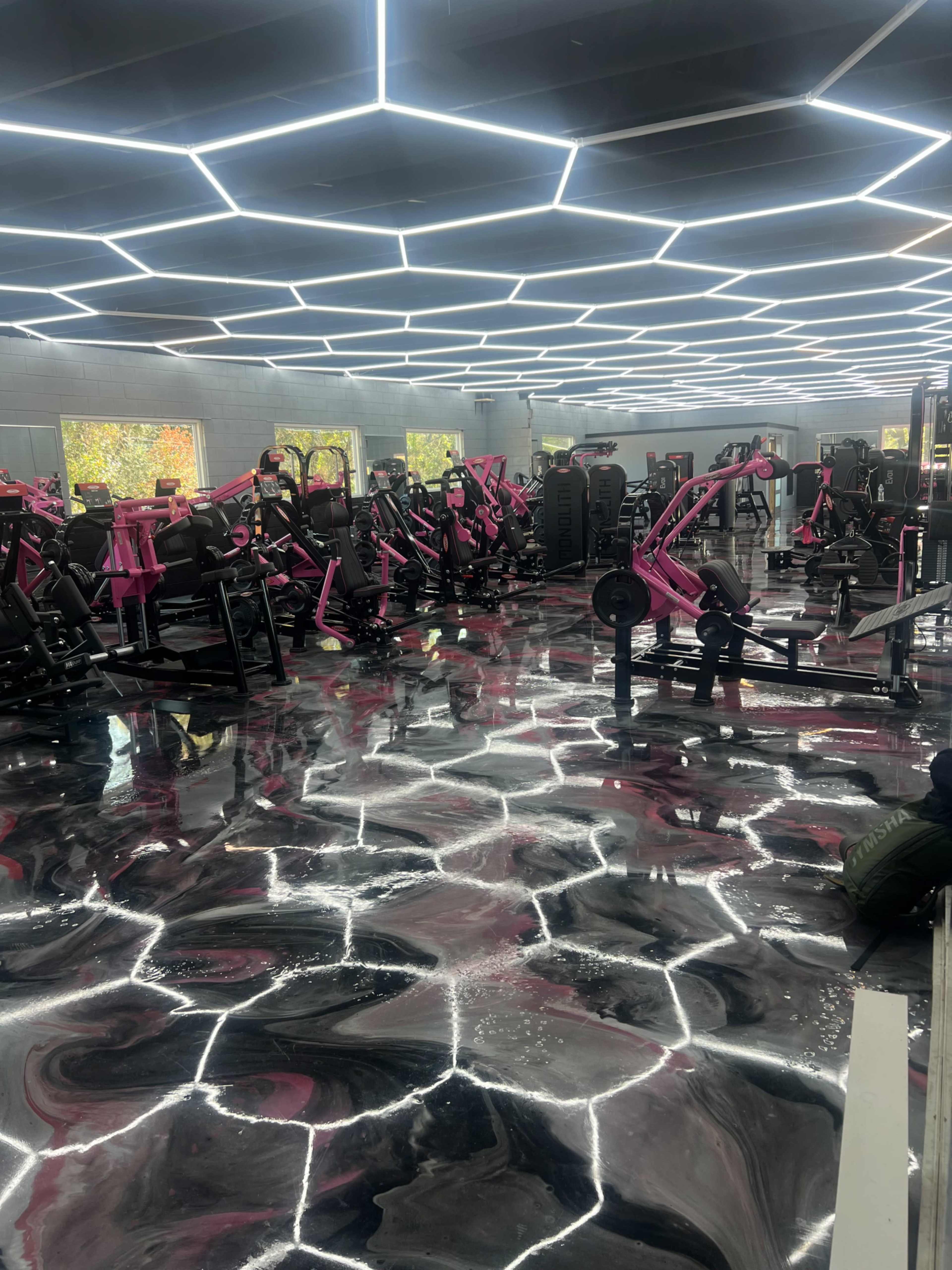 The image shows a modern gym interior featuring an array of fitness equipment in shades of pink, complemented by a glossy black and white floor and bright geometric lighting.