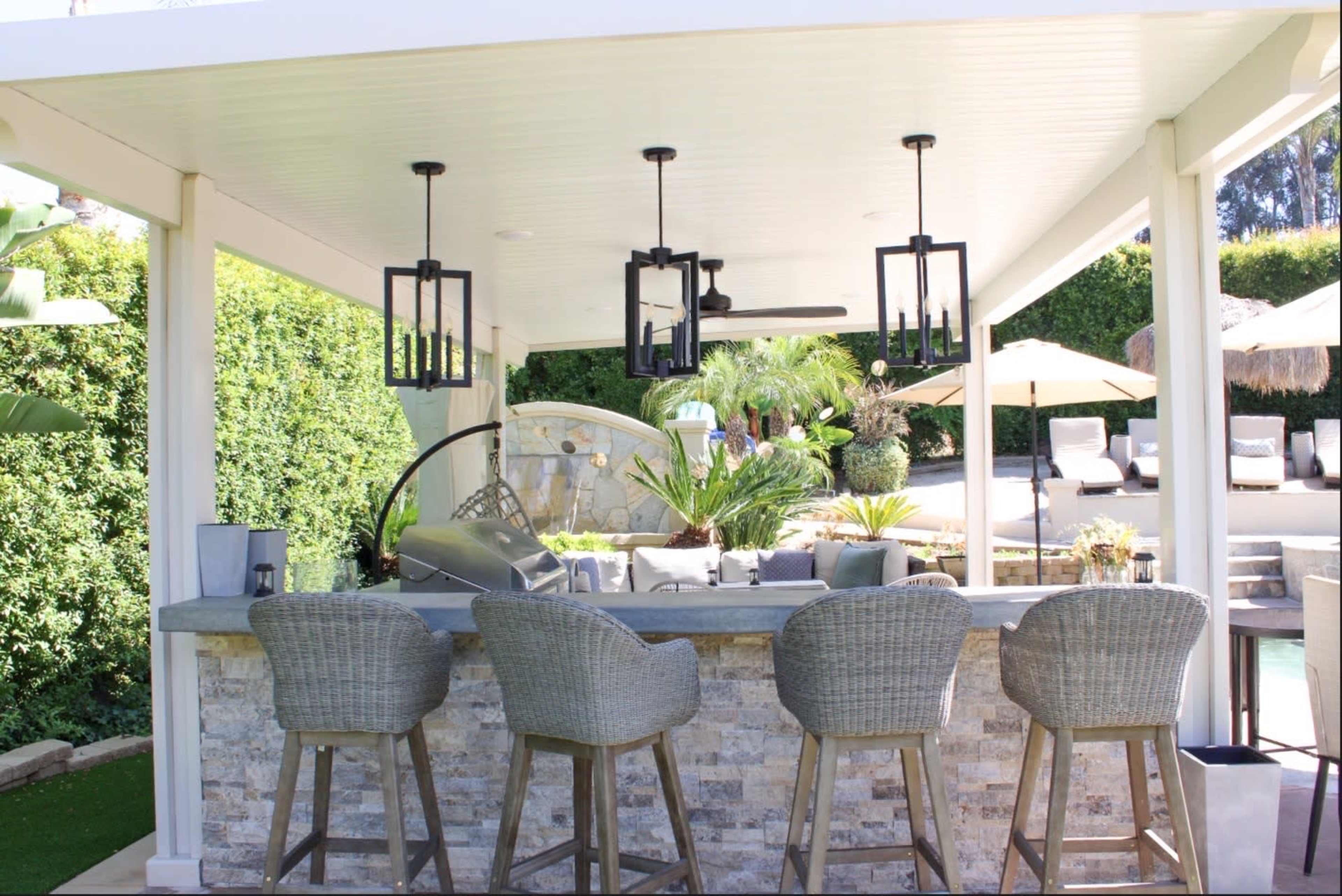 The image shows an outdoor bar area with stone detailing, four woven bar stools, and pendant lighting under a covered roof.