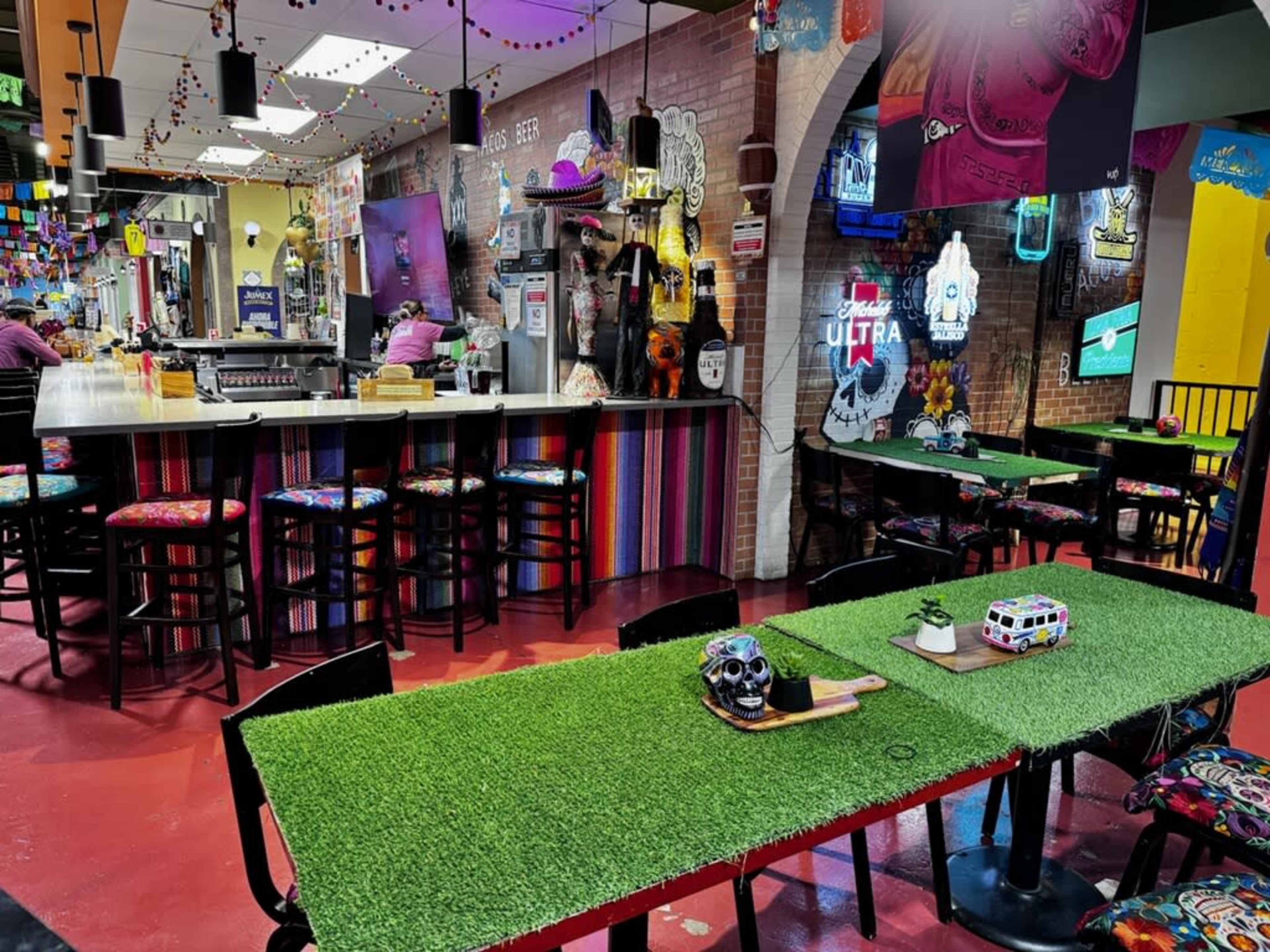 The image shows a vibrant restaurant interior with colorful chairs, artificial grass-covered tables, and a bar area in the background adorned with eclectic decorations.