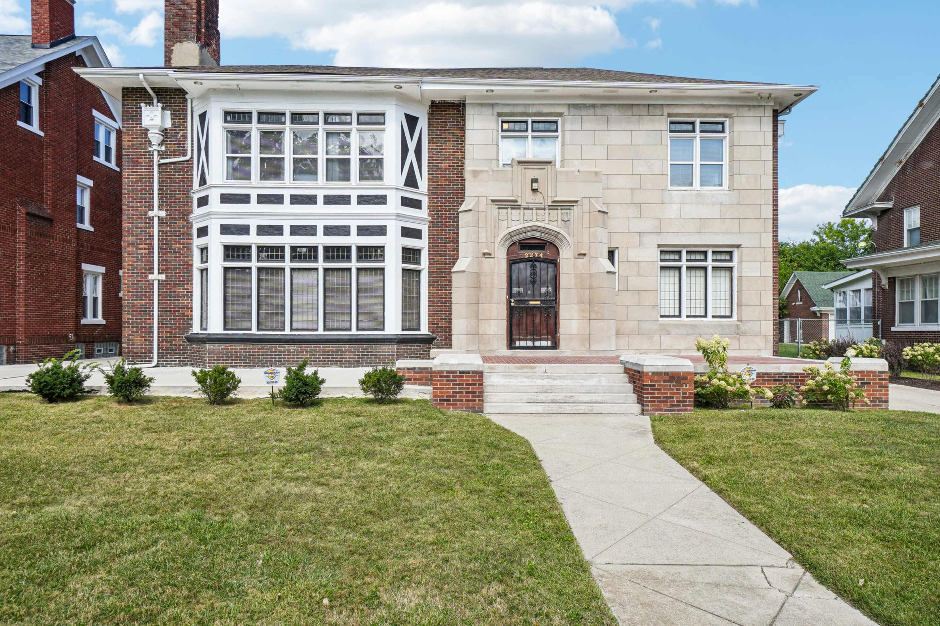 The image shows a large, two-story house with a combination of stone and brick exterior, featuring large windows and decorative architectural elements.