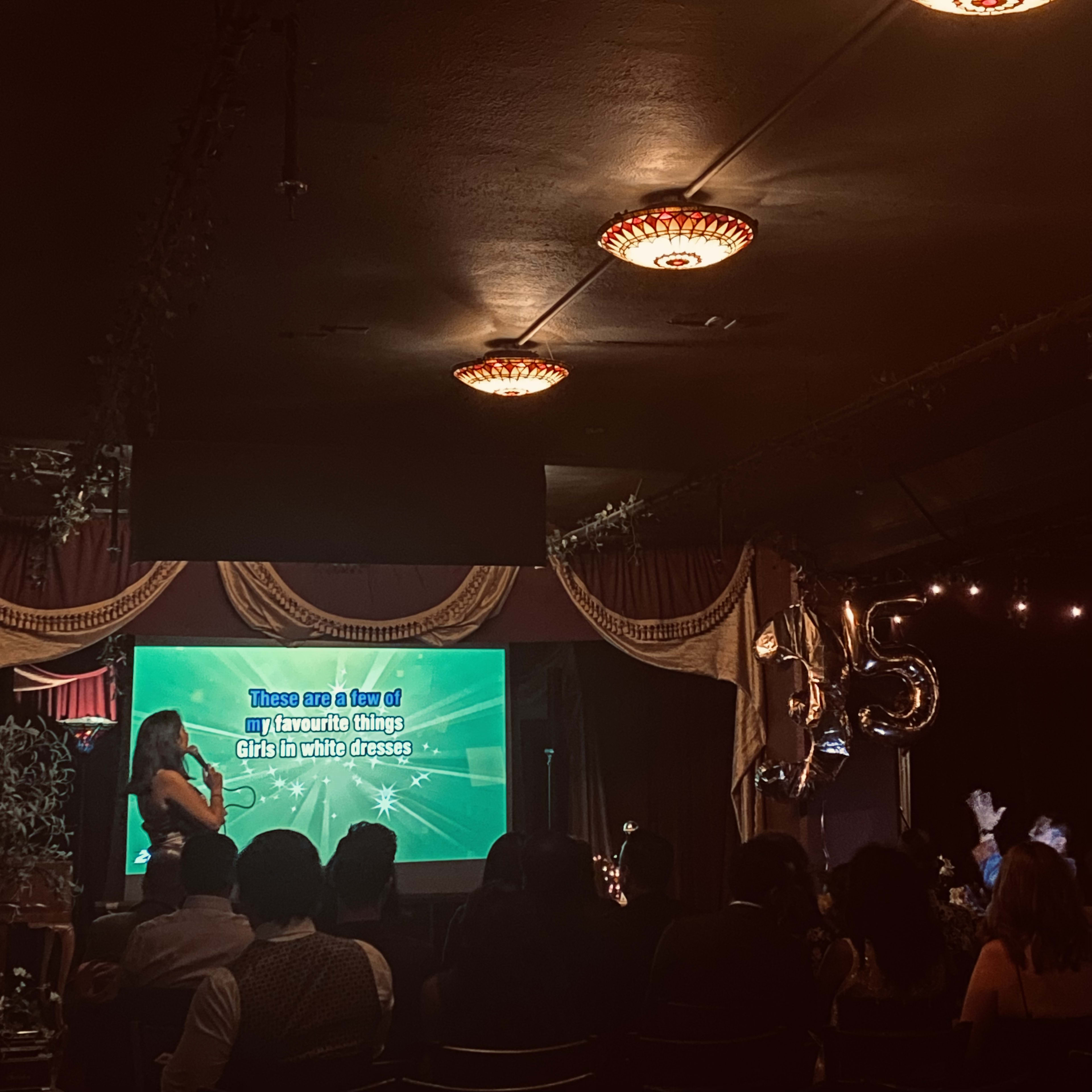 A performer stands on stage in front of a screen displaying lyrics, while the audience watches in a dimly lit venue.