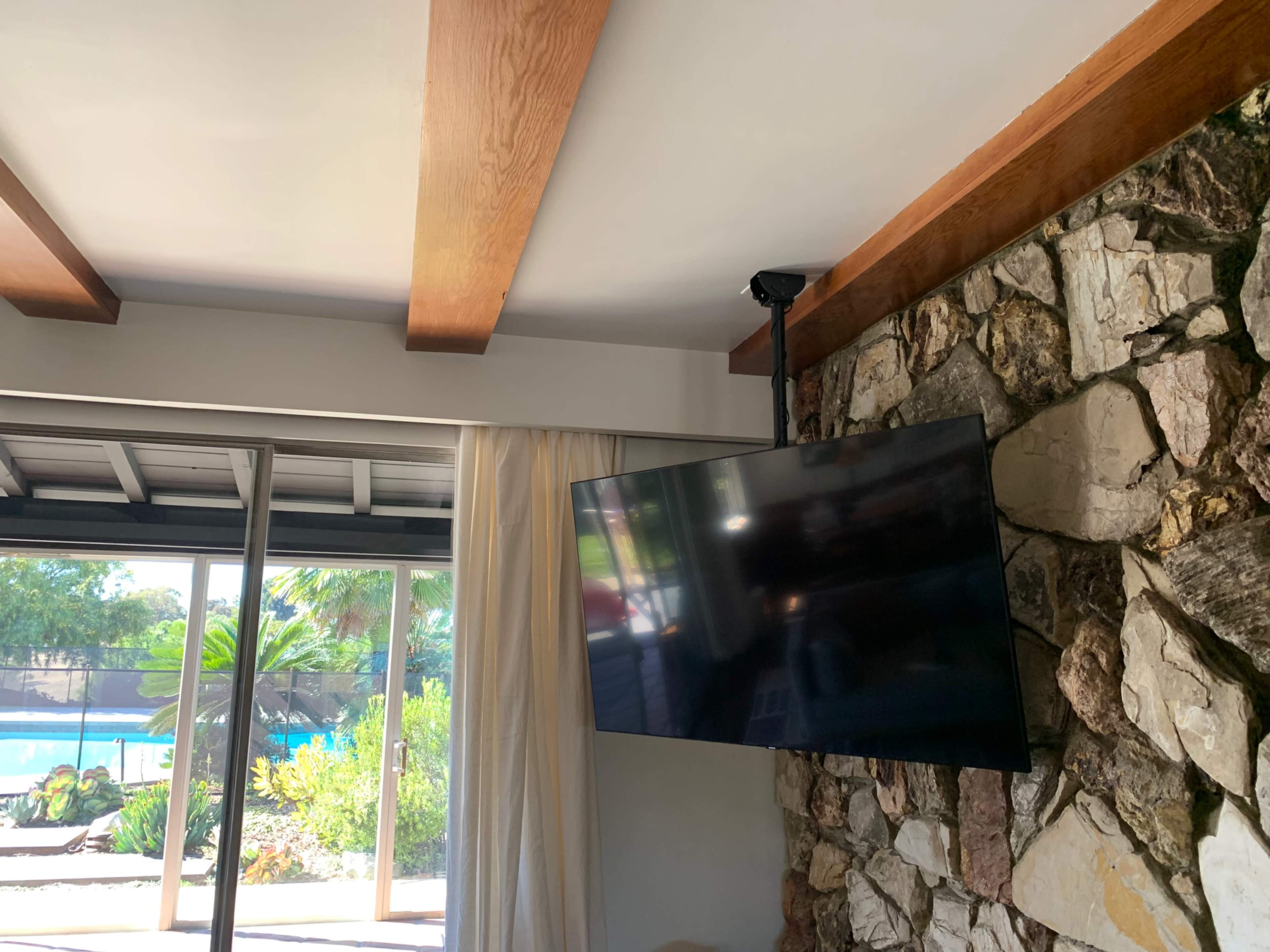 A flat-screen television is mounted on a stone wall with wooden beams above in a room with large glass doors leading to an outdoor area.