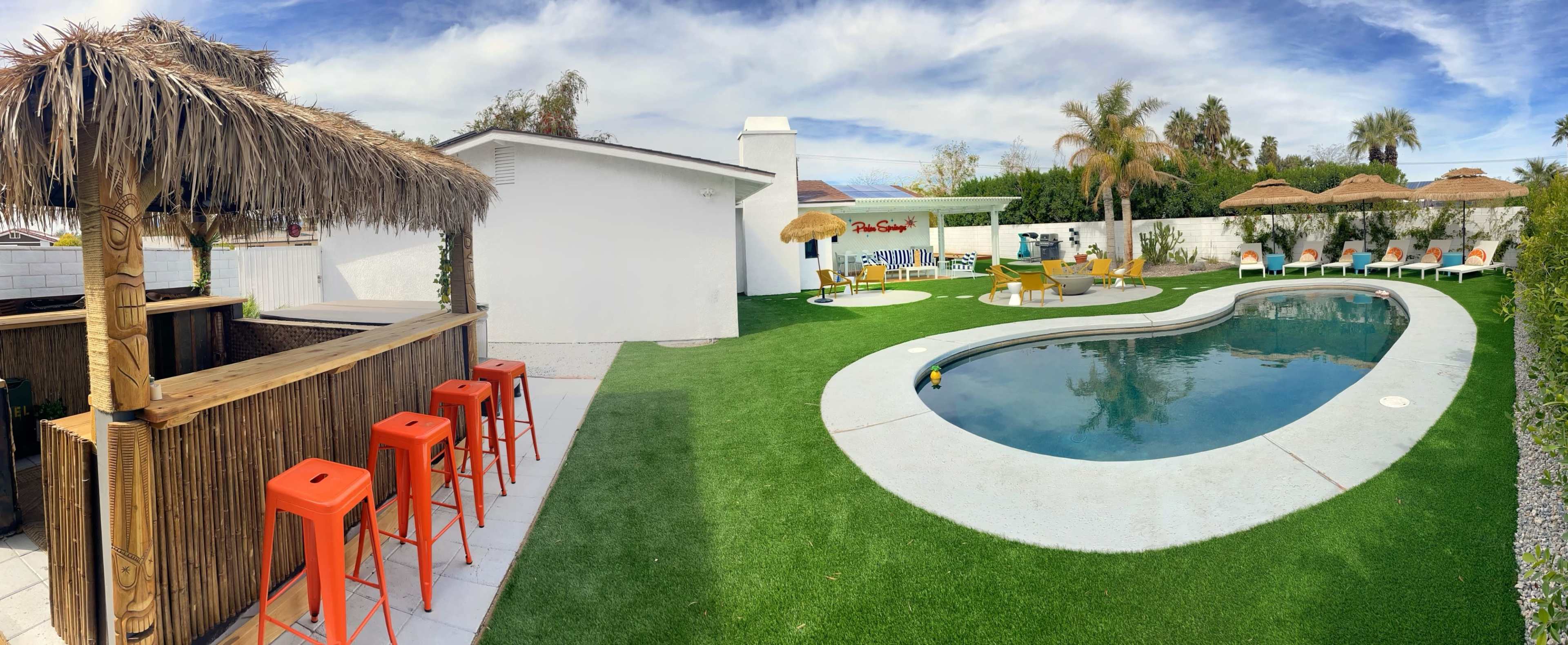 The image shows a landscaped outdoor area featuring a swimming pool, a tiki bar with orange stools, lounge chairs, and several palm trees under a blue sky.