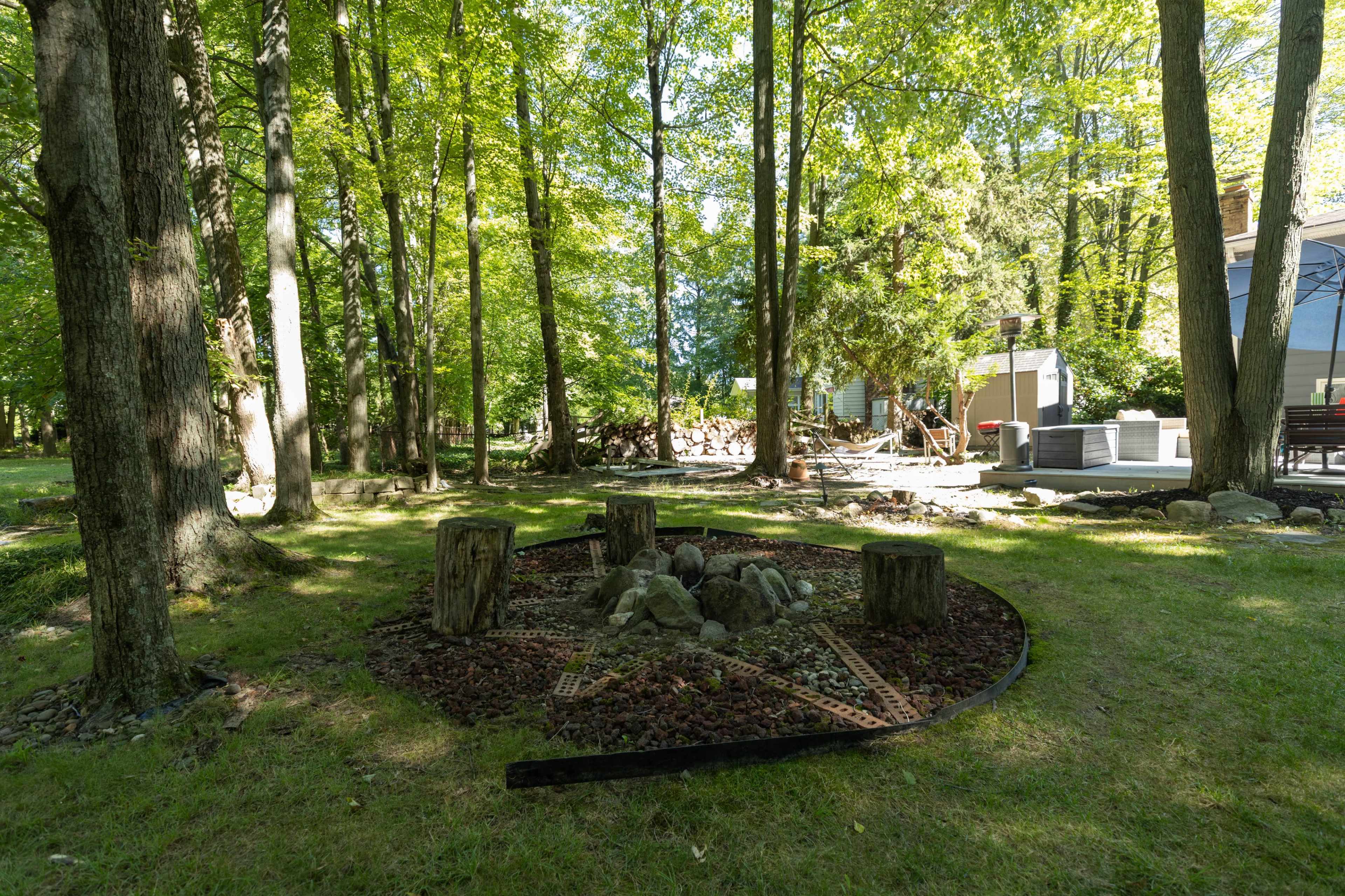 The image shows a circular fire pit area with wooden stumps arranged around it, surrounded by trees and a residential building in the background.