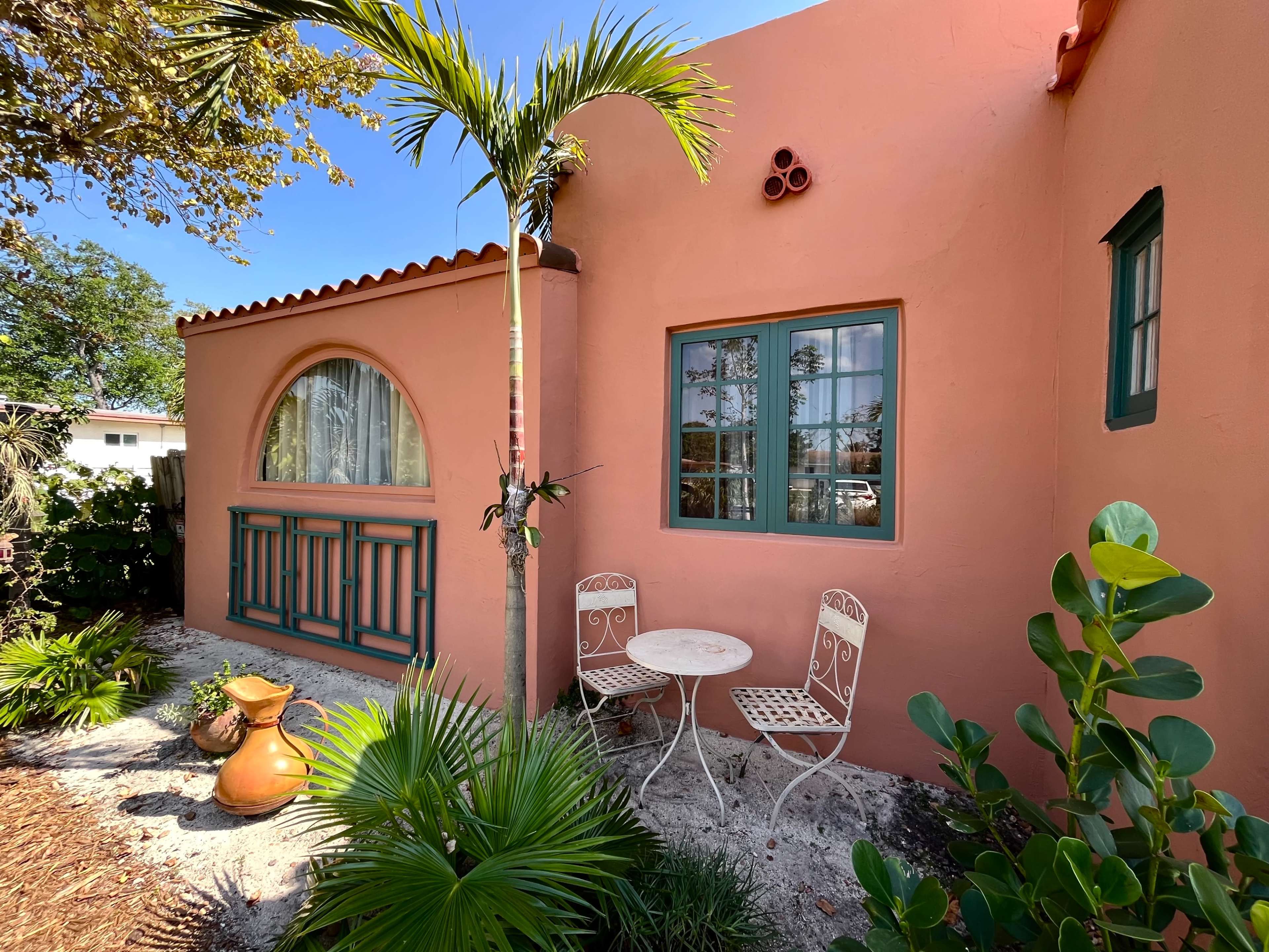 A small patio with two white metal chairs and a table sits next to a pink stucco building, surrounded by lush greenery and a terracotta pot.
