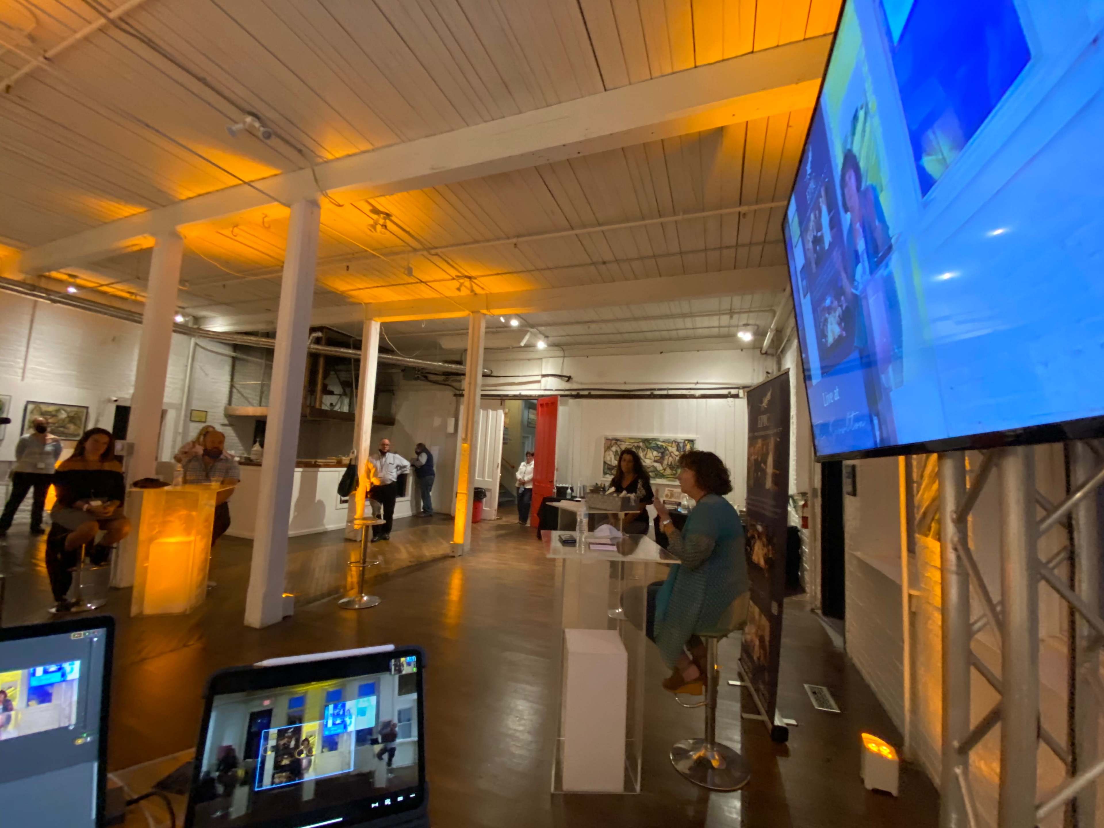 A spacious indoor event with people mingling, a presentation taking place on a large screen, and illuminated columns enhancing the setting.