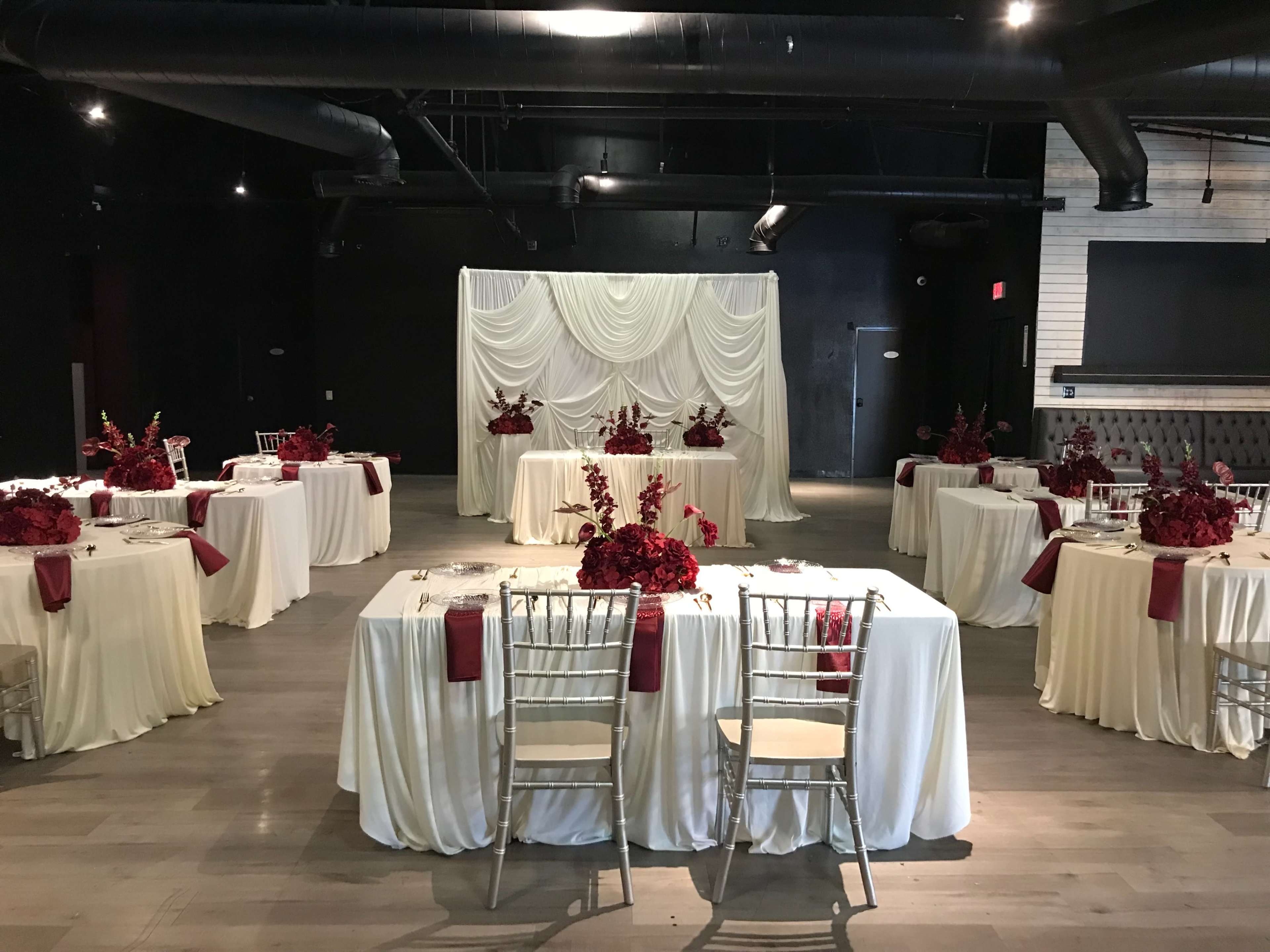 The image shows a spacious event venue arranged with multiple elegantly set dining tables adorned with white tablecloths and red floral centerpieces.