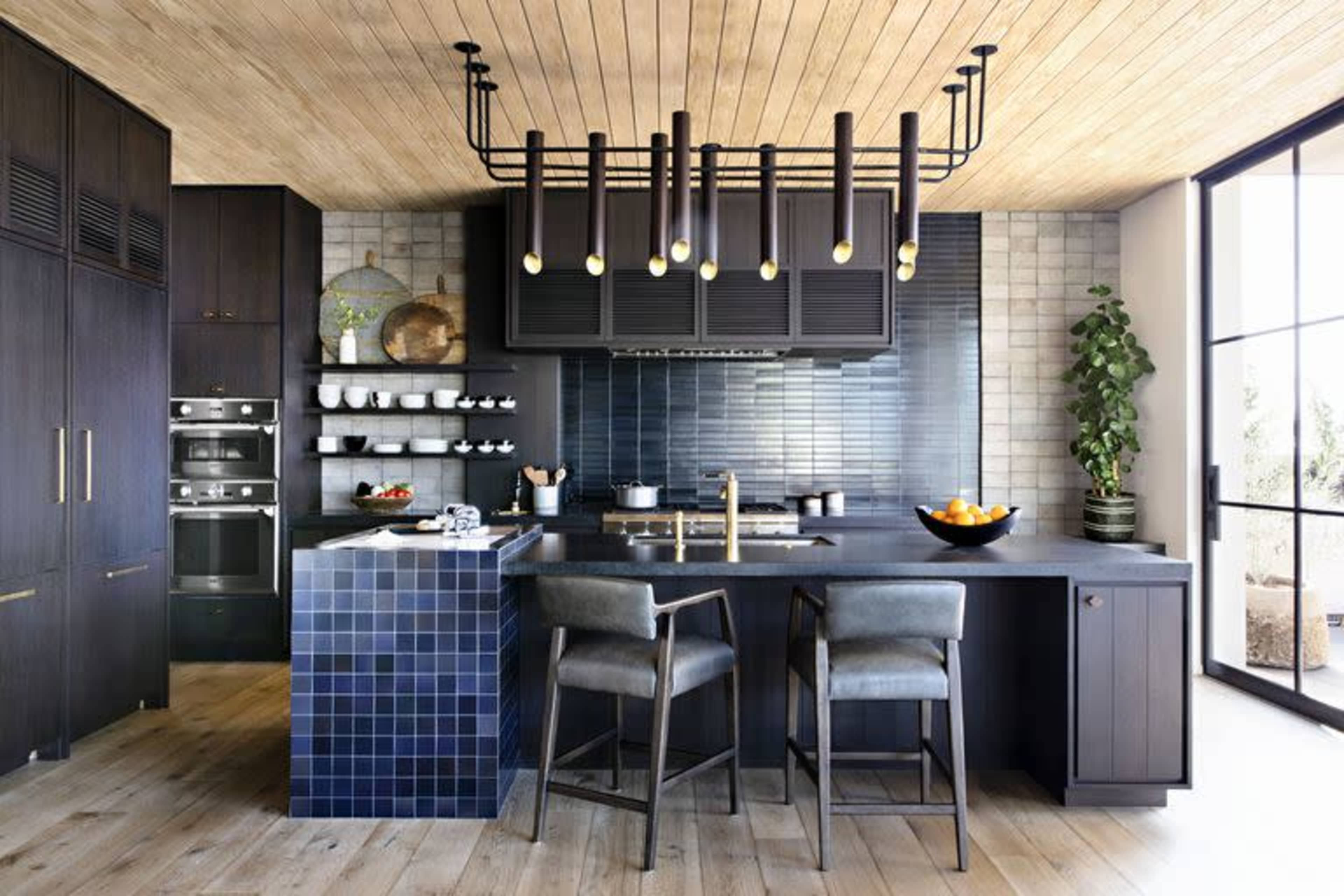 A modern kitchen features dark cabinetry, a tiled backsplash, and a central island with bar stools, illuminated by pendant lights.