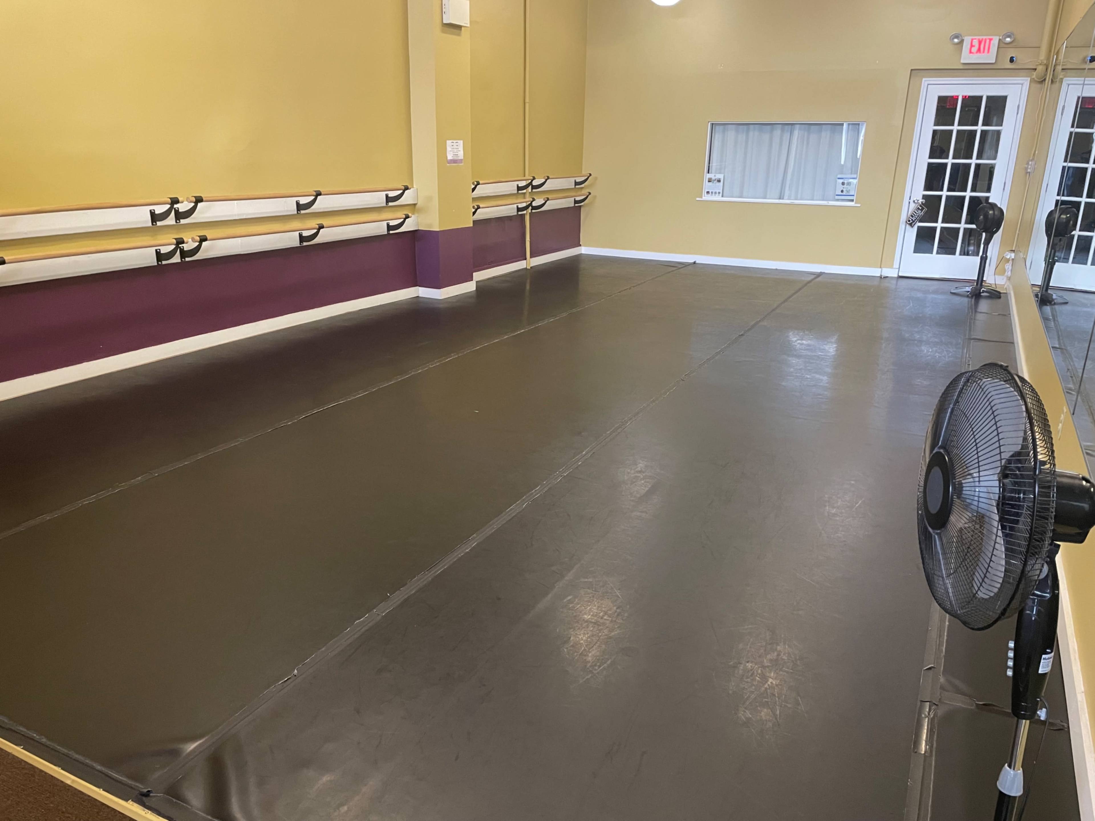 The image shows an empty dance studio with black flooring, a fan on the side, and walls painted in yellow and purple.