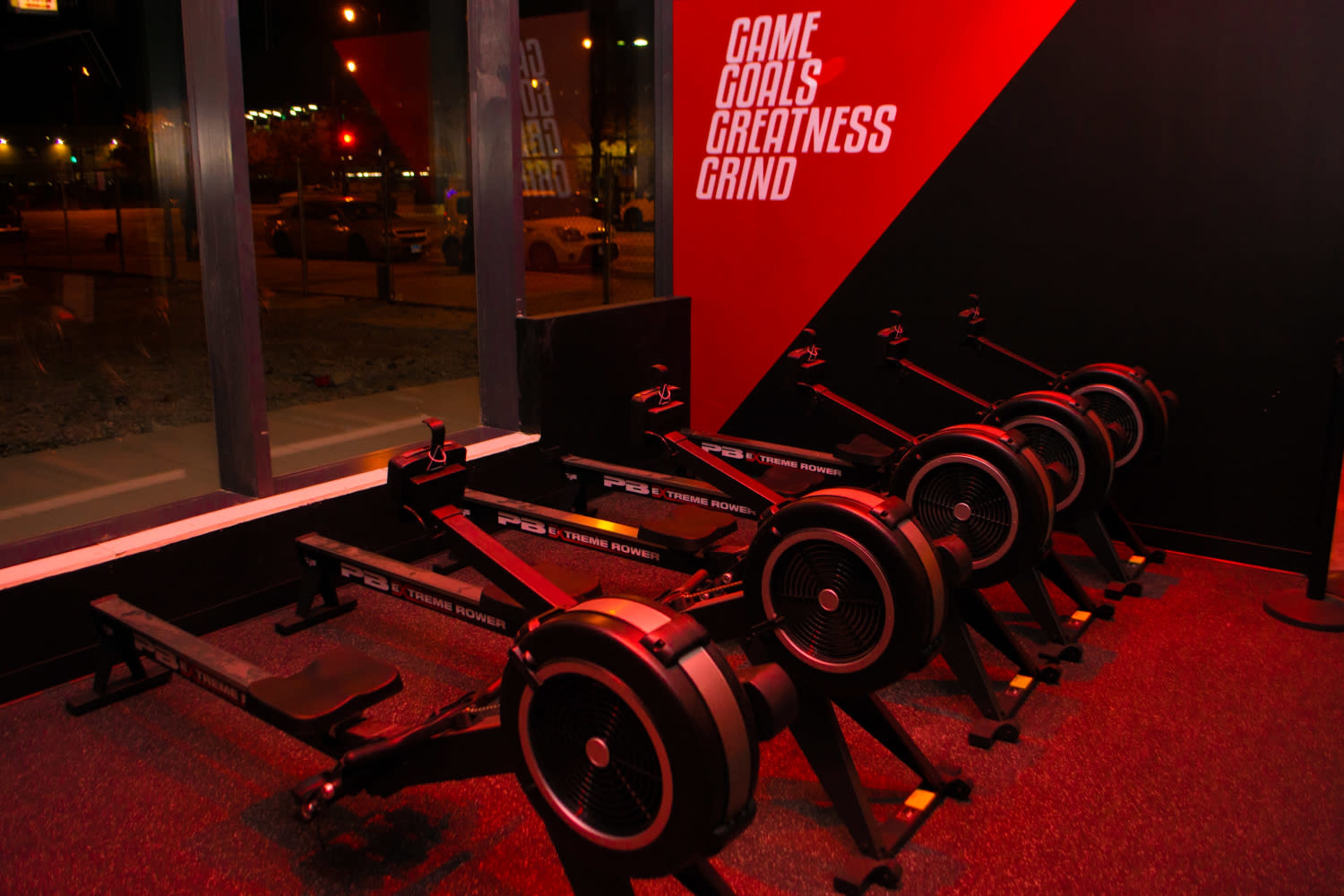 A row of four indoor rowing machines is positioned against a wall with motivational phrases in a dimly lit gym space.