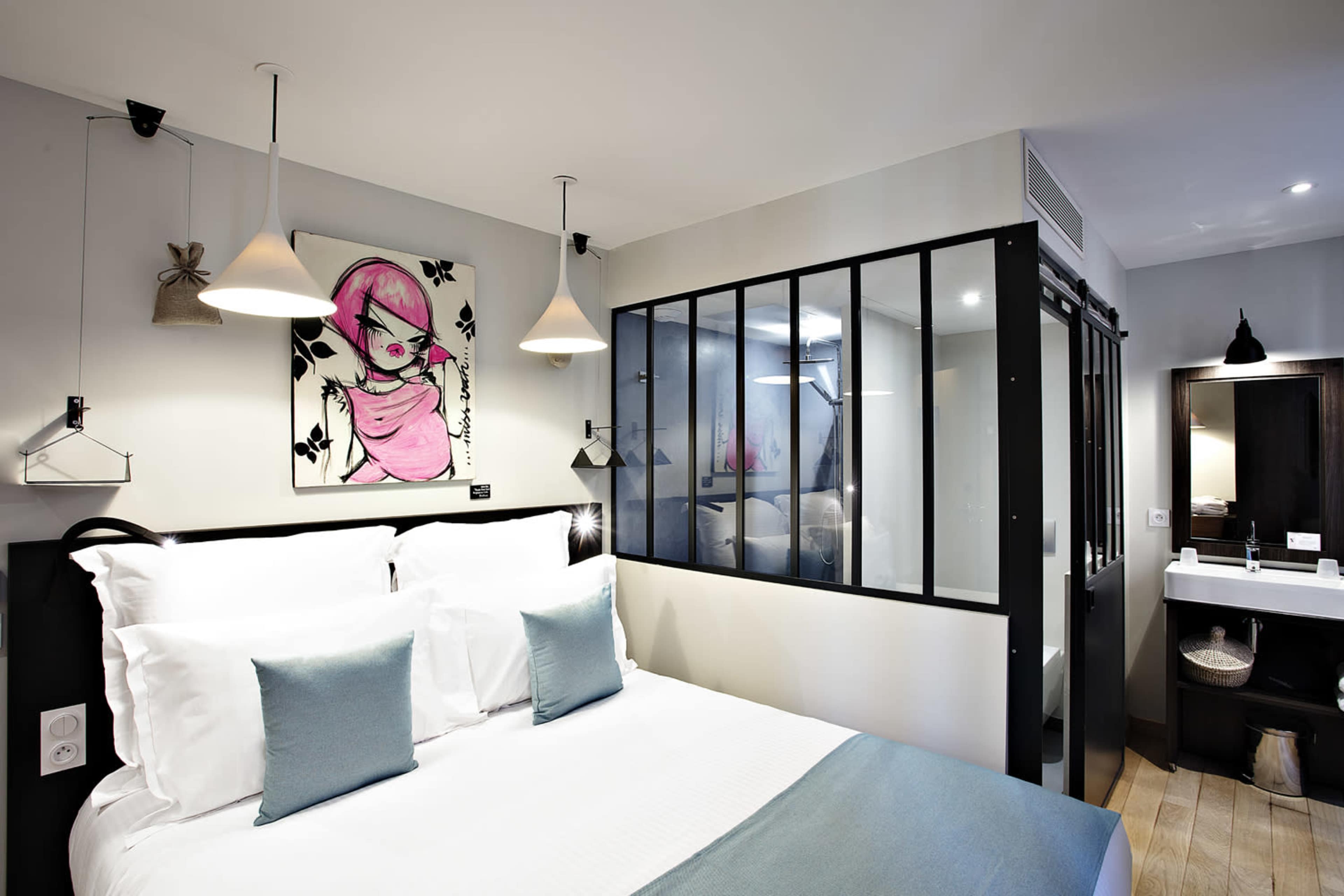 The image shows a modern hotel room featuring a double bed with decorative pillows, a glass partitioned shower area, and artistic wall decor.