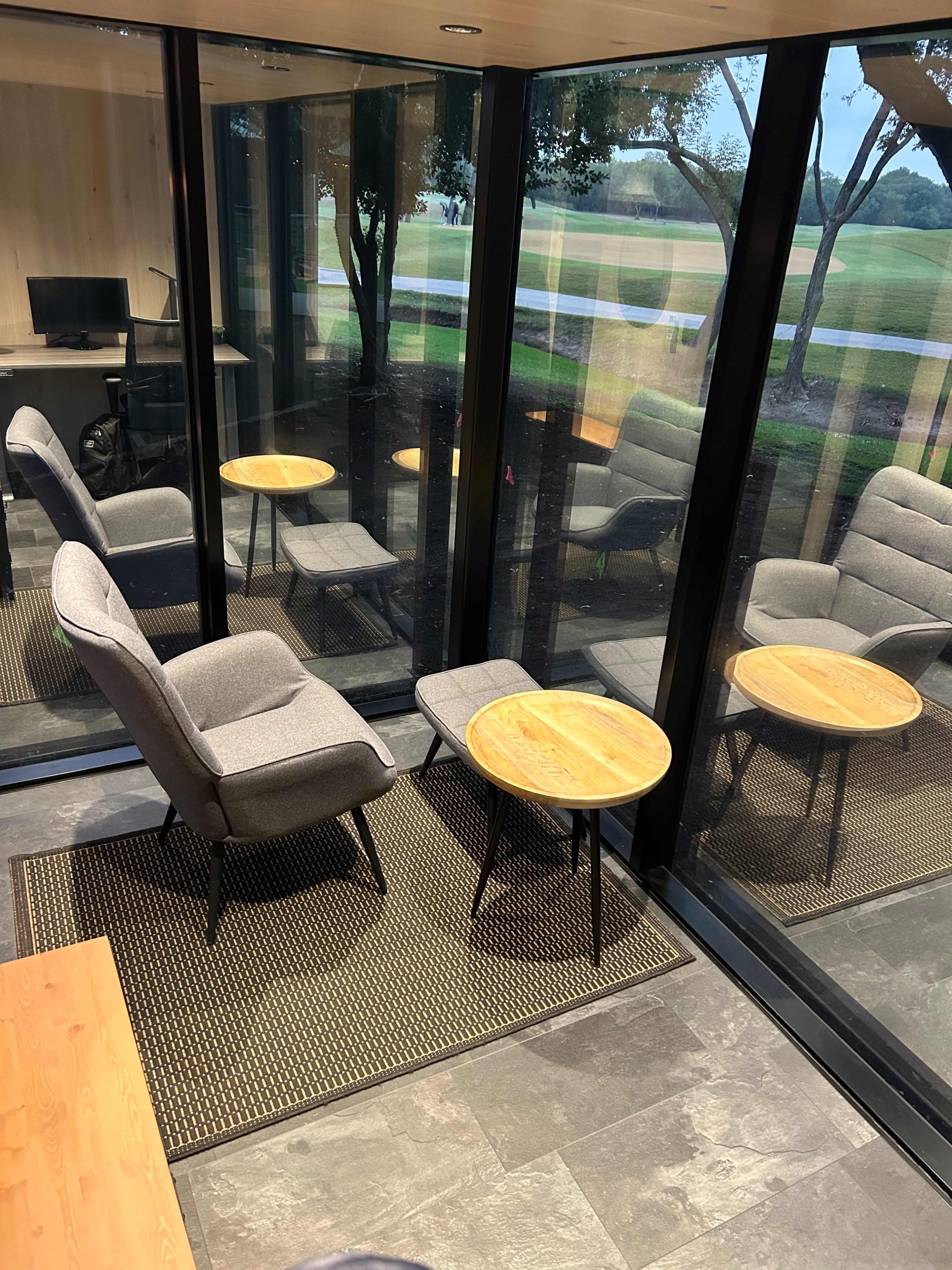 A modern glass-walled room with two gray armchairs and a wooden side table on a dark tiled floor, overlooking a grassy area outside.