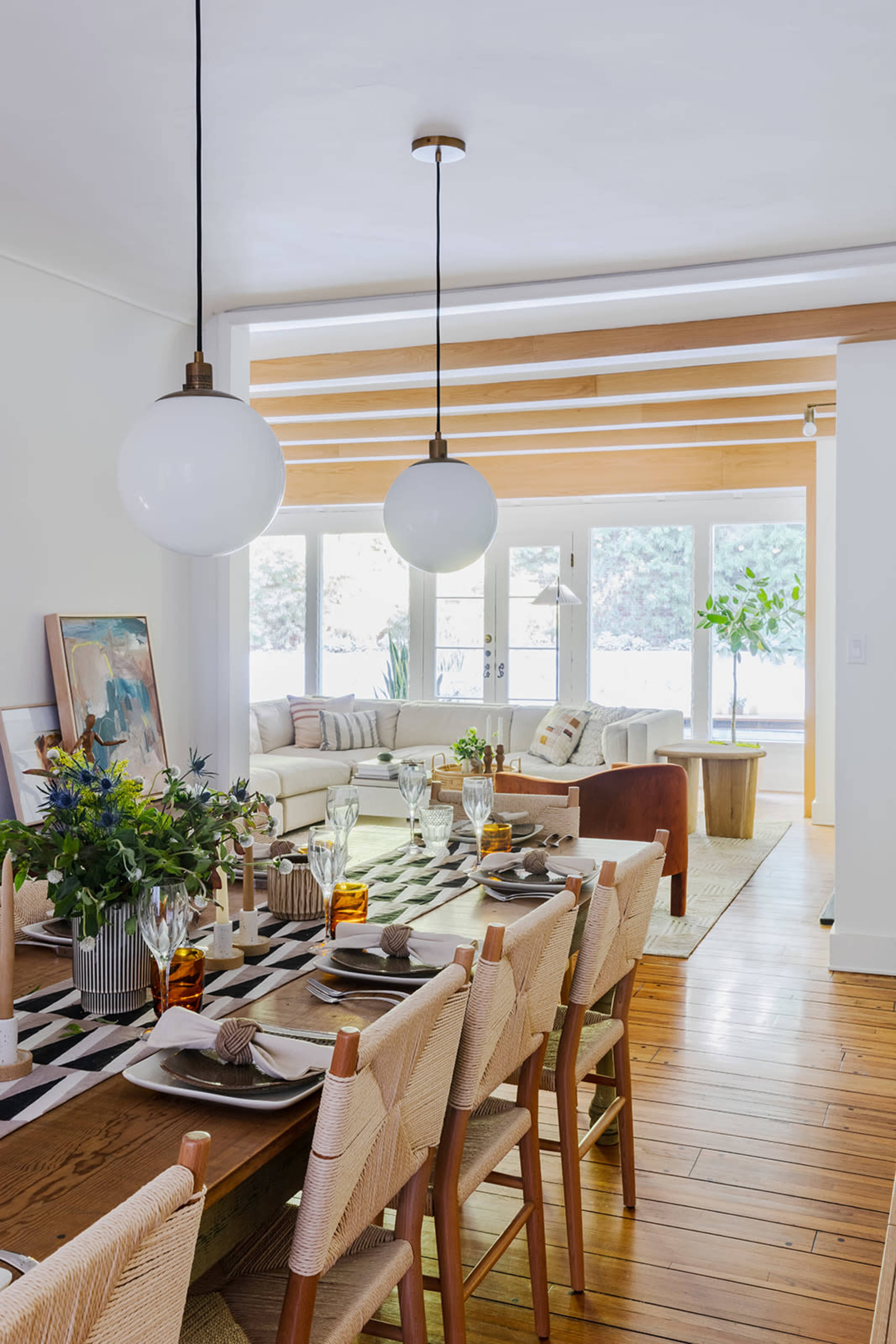 A modern dining area features a long table set with dishes and glasses, illuminated by hanging spherical lights, surrounded by a cozy living space with a sectional sofa and large windows.