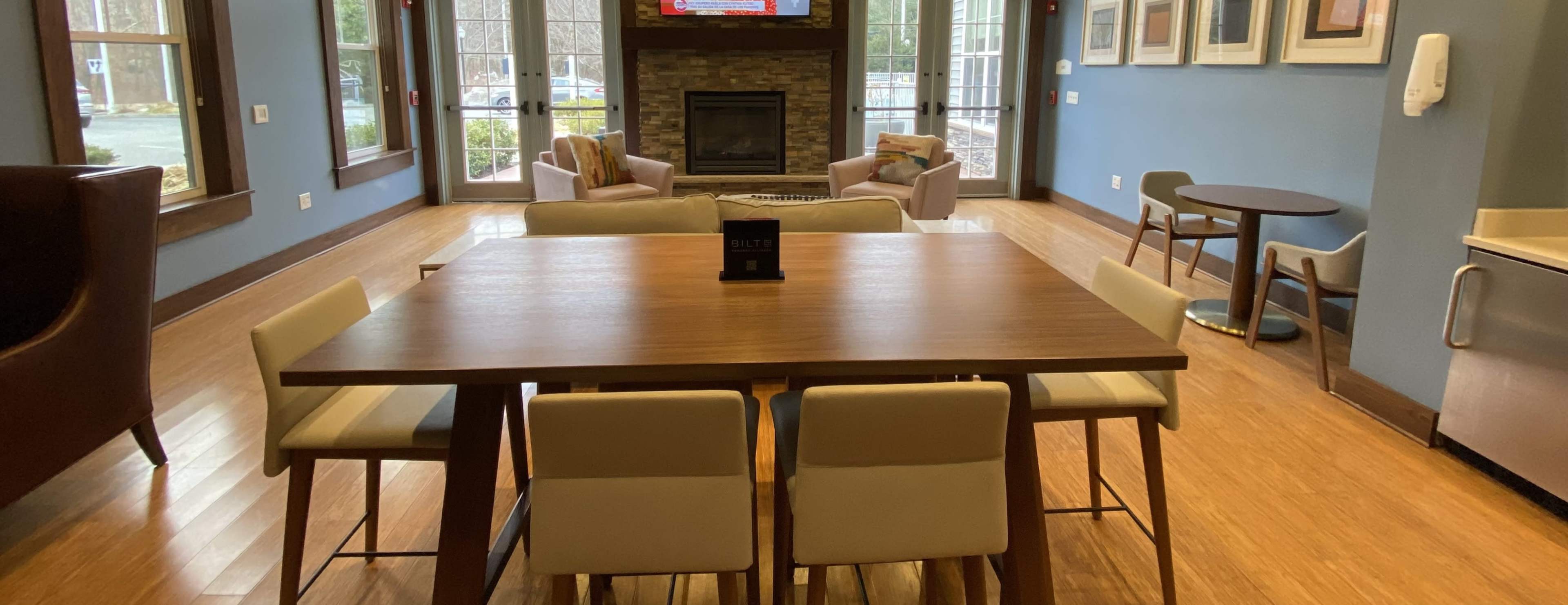 A communal lounge area featuring a central wooden table surrounded by white chairs, with a fireplace and seating arrangement visible in the background.