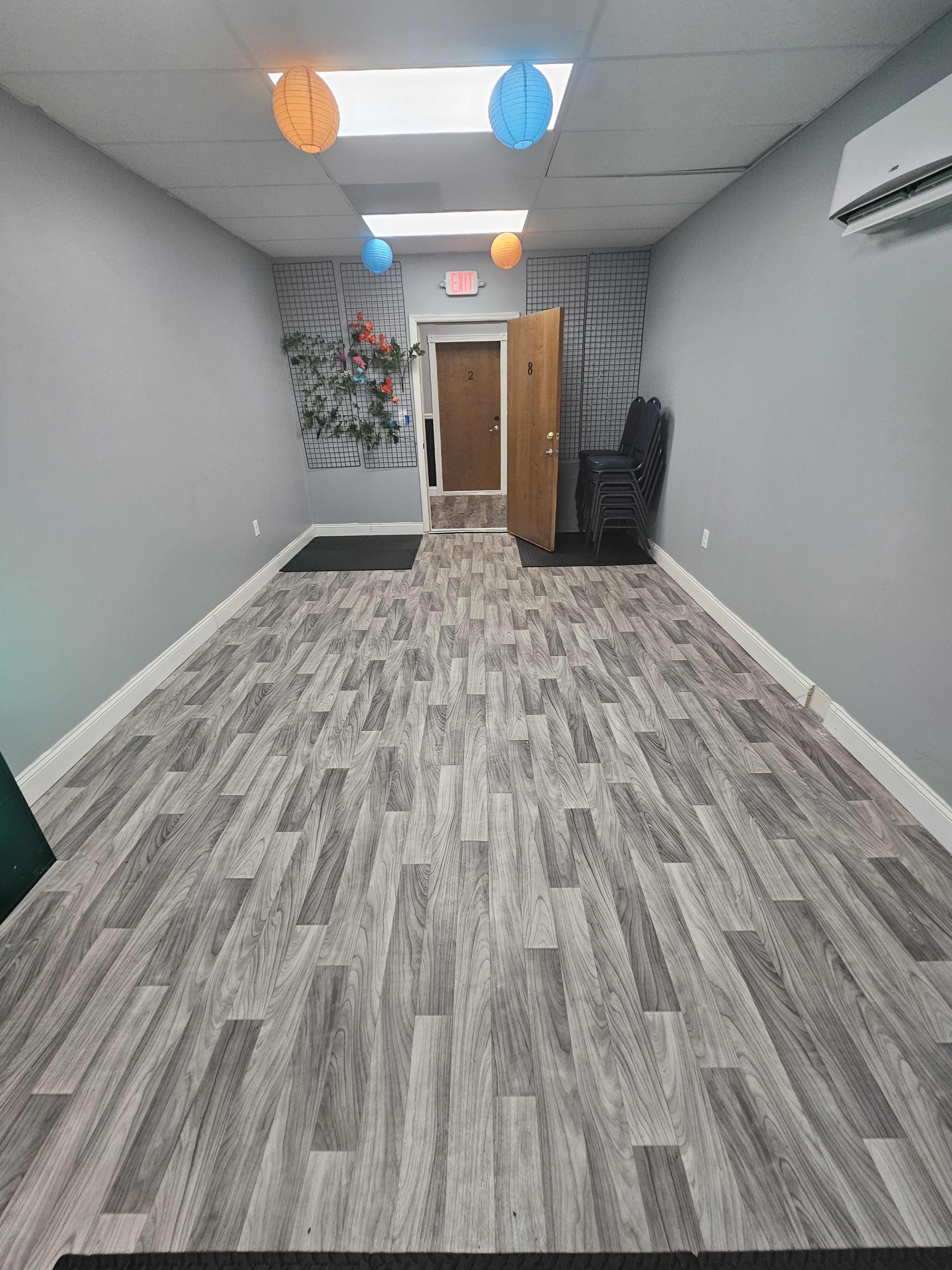 A narrow hallway with light gray wood-like flooring, a door at the end, and colorful lanterns hanging from the ceiling.