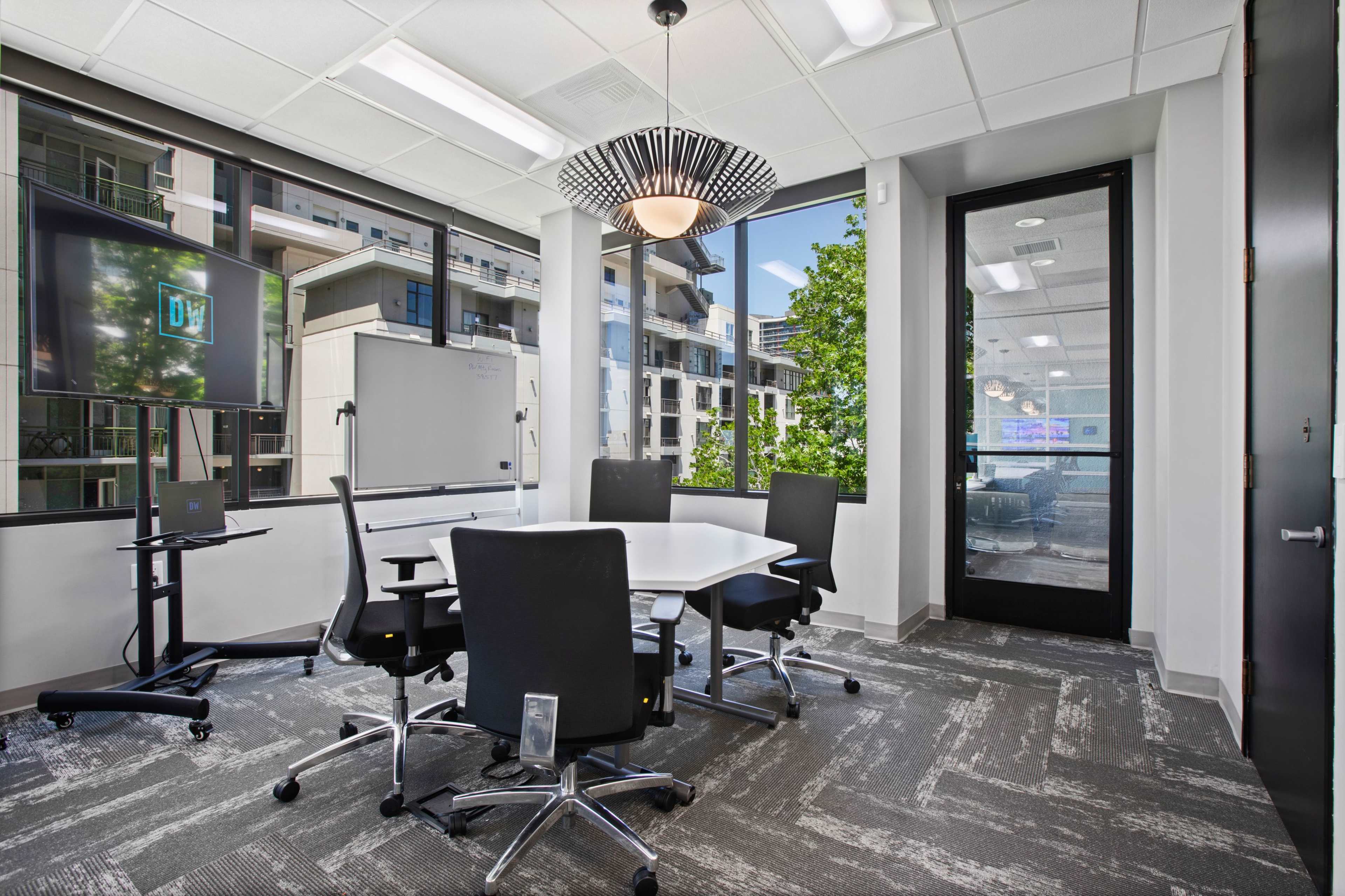 A modern conference room features a white table surrounded by black chairs, large windows showing an outdoor view, and equipped with a TV and whiteboard.