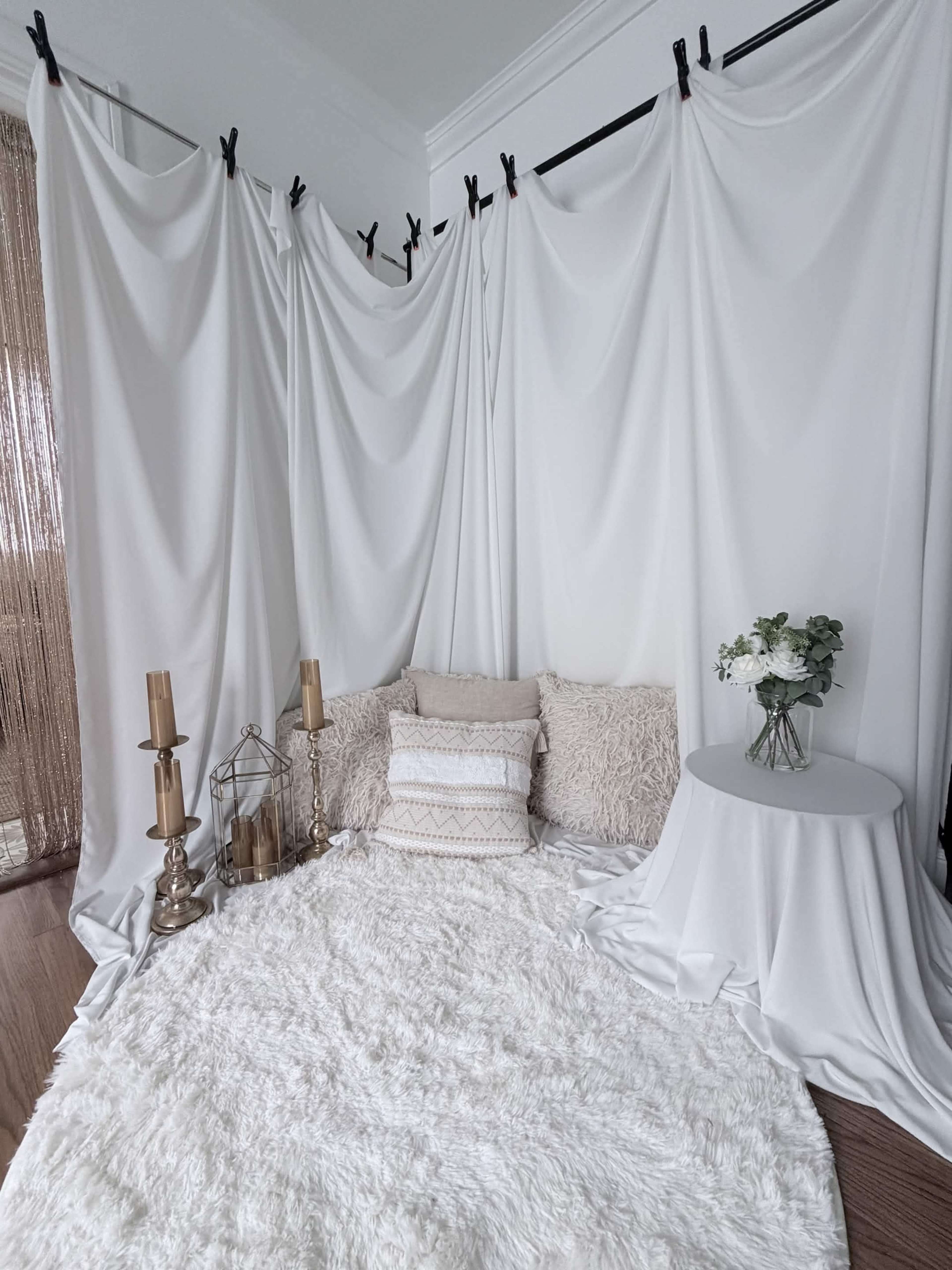 The scene features a cozy, white-themed indoor space with draped fabric backgrounds, a plush rug, decorative pillows, candles, and a small table adorned with flowers.