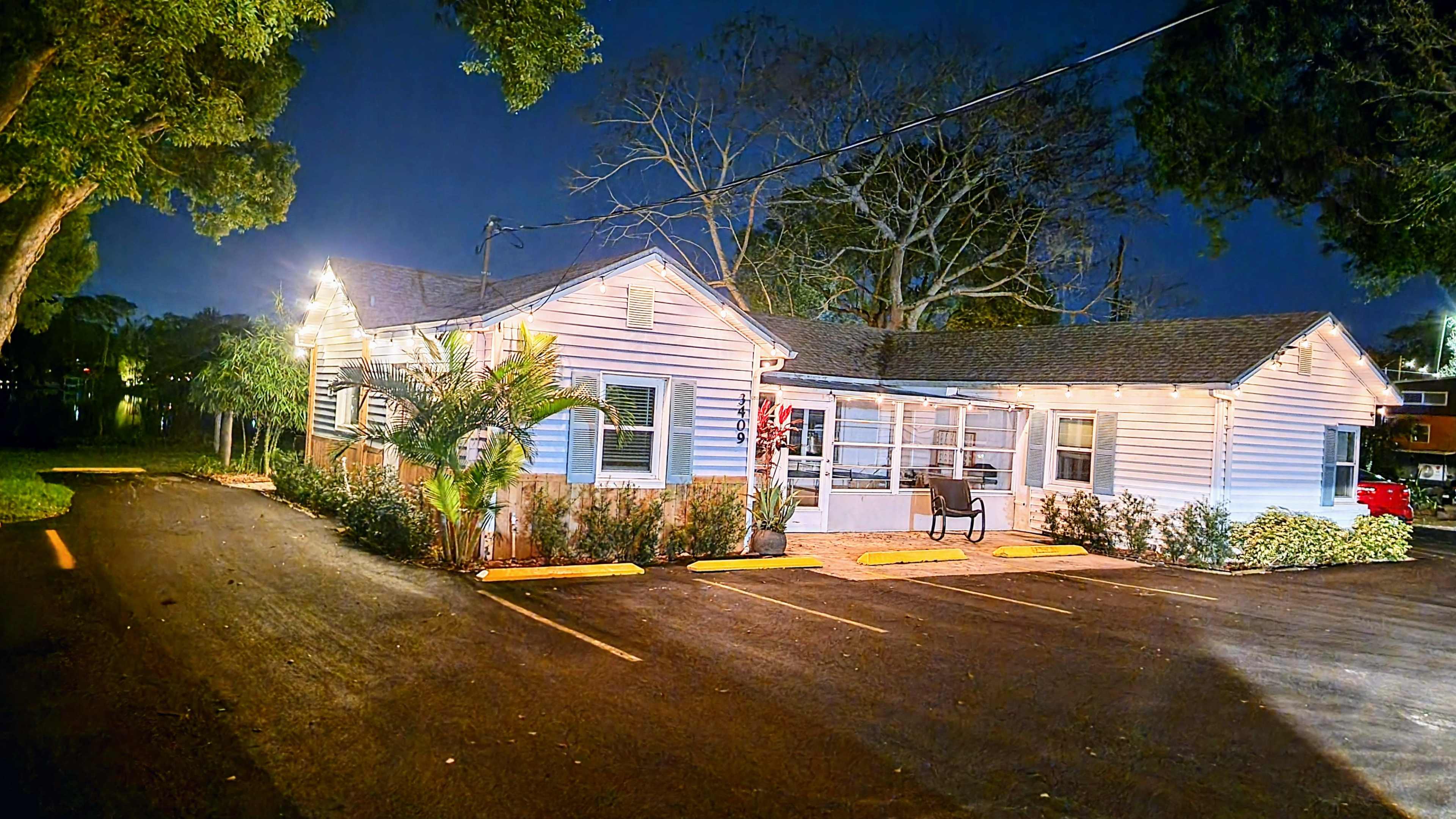 A single-story, pink house with a porch is illuminated by string lights, surrounded by trees and parked cars on a dark street.
