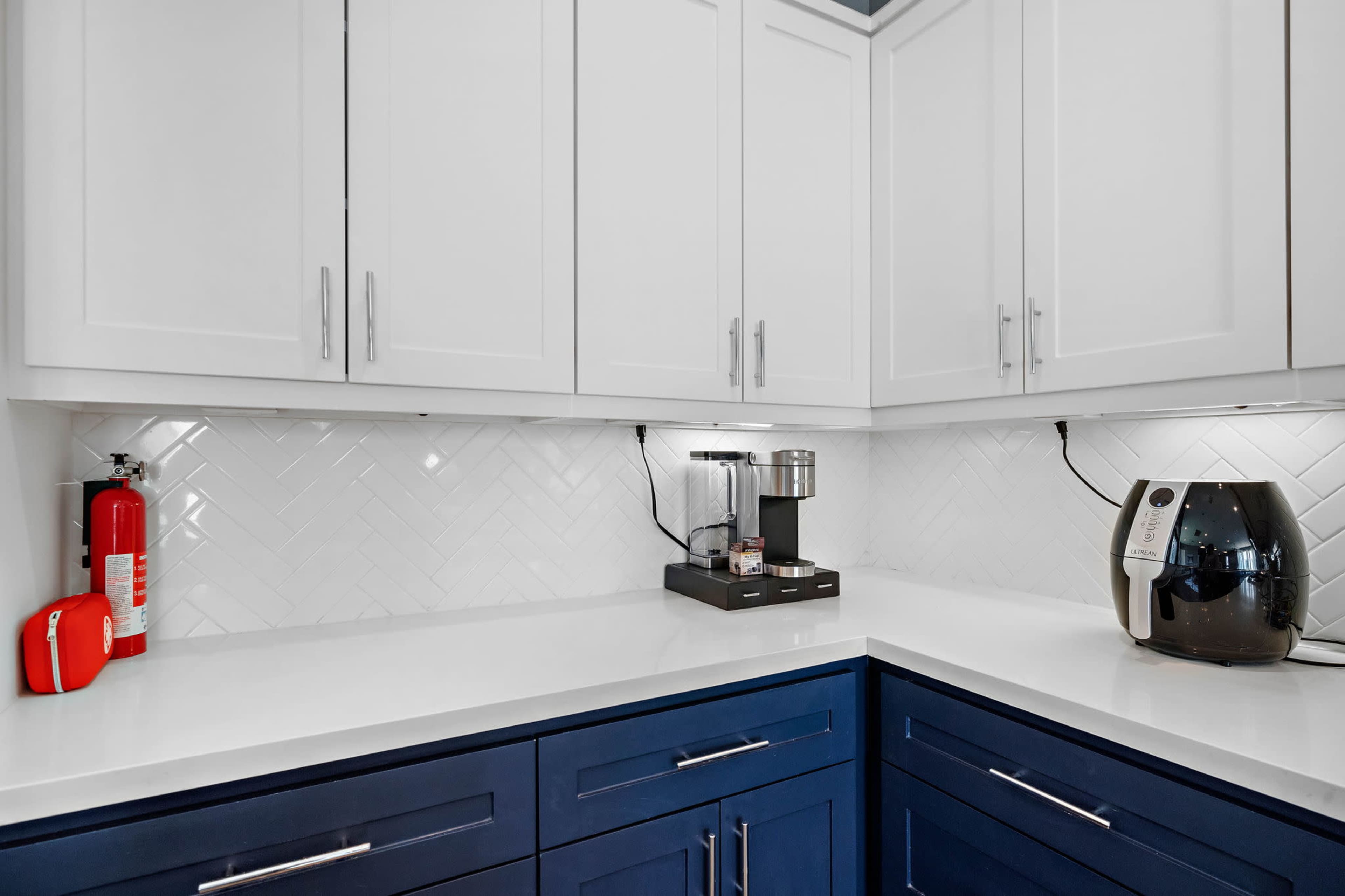 A modern kitchen corner featuring blue cabinets, a coffee maker, an air fryer, and a fire extinguisher against a white backsplash with herringbone patterns.