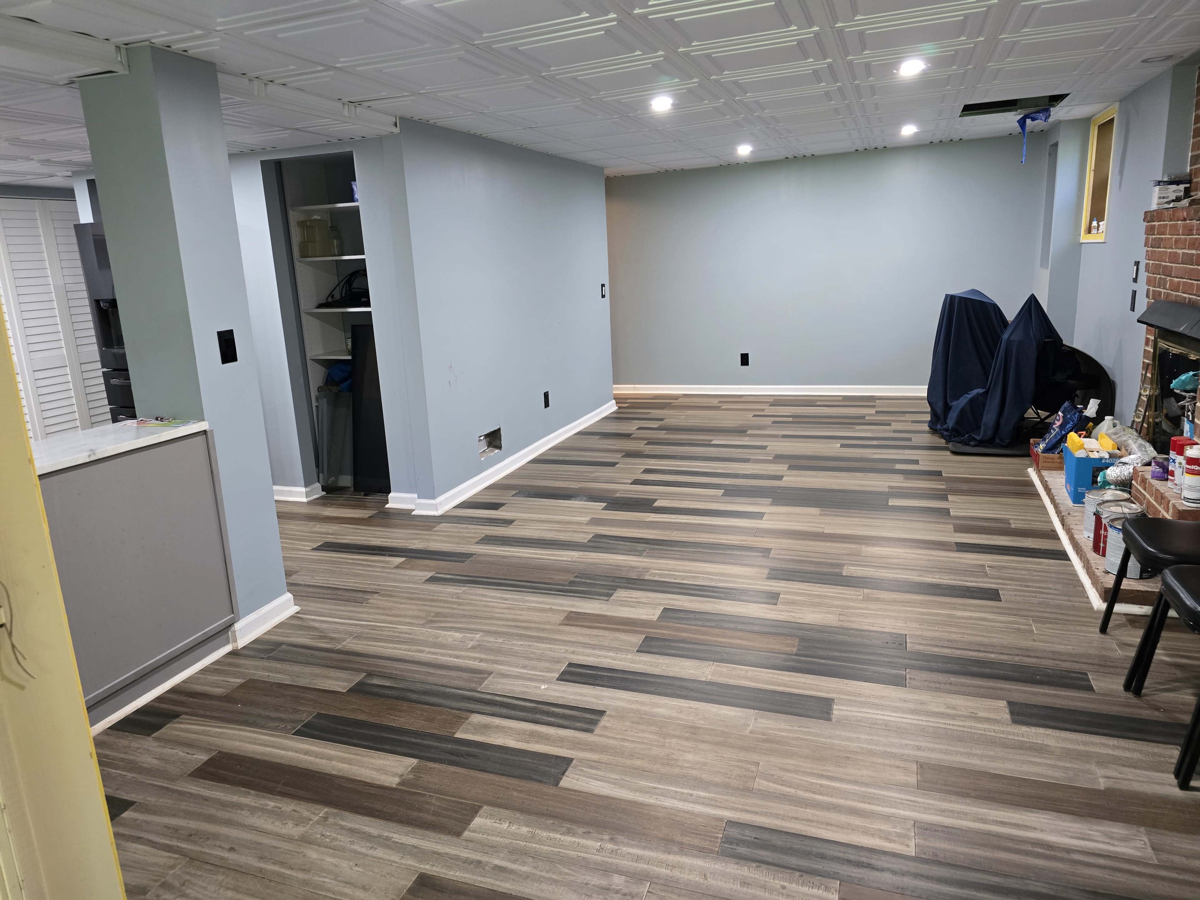 The image shows a vacant basement with newly installed laminate flooring, light blue walls, and an unfinished area with a fireplace and some scattered tools and materials.