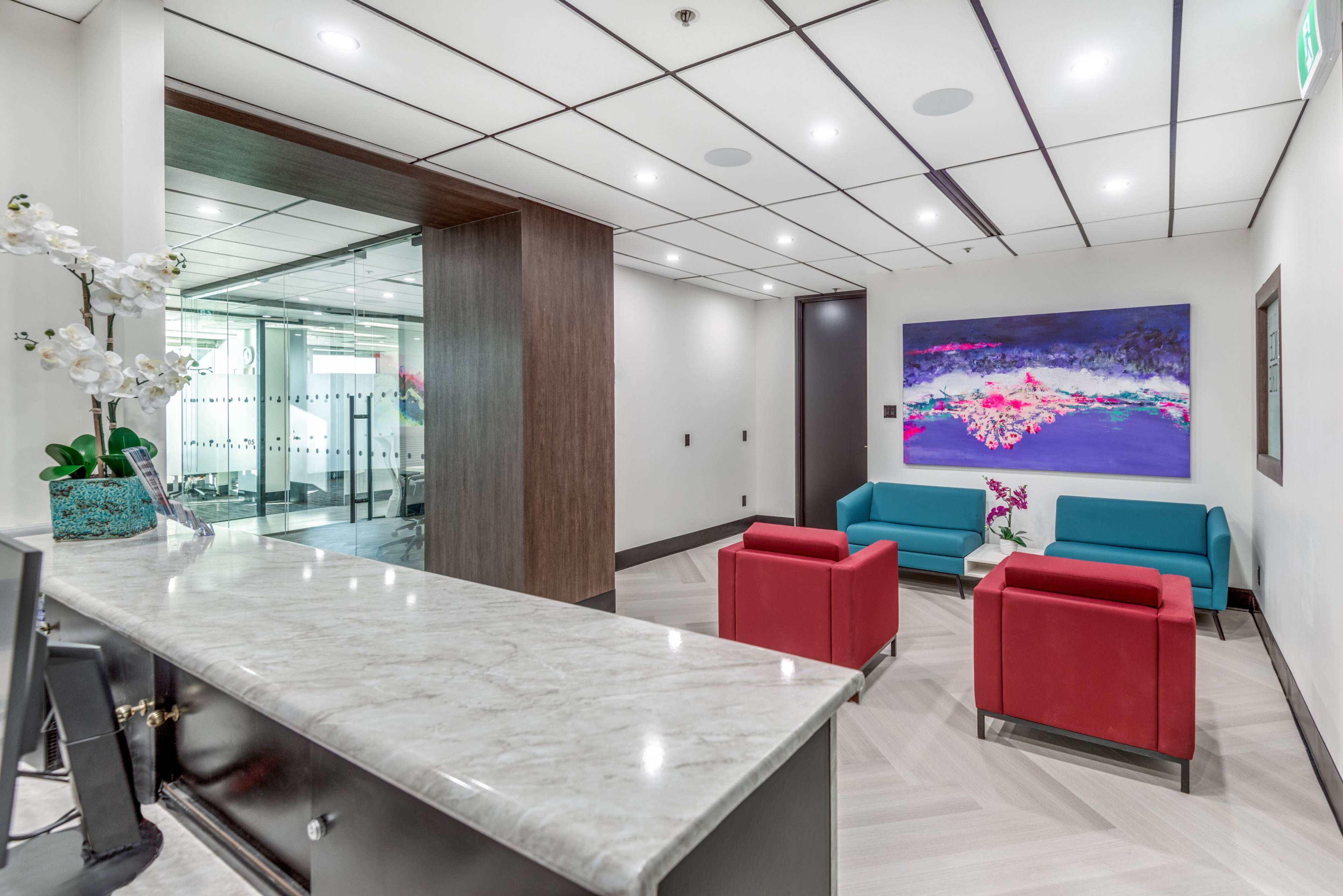 A modern reception area with a marble counter, two teal sofas, and two red armchairs, complemented by a colorful abstract painting on the wall.