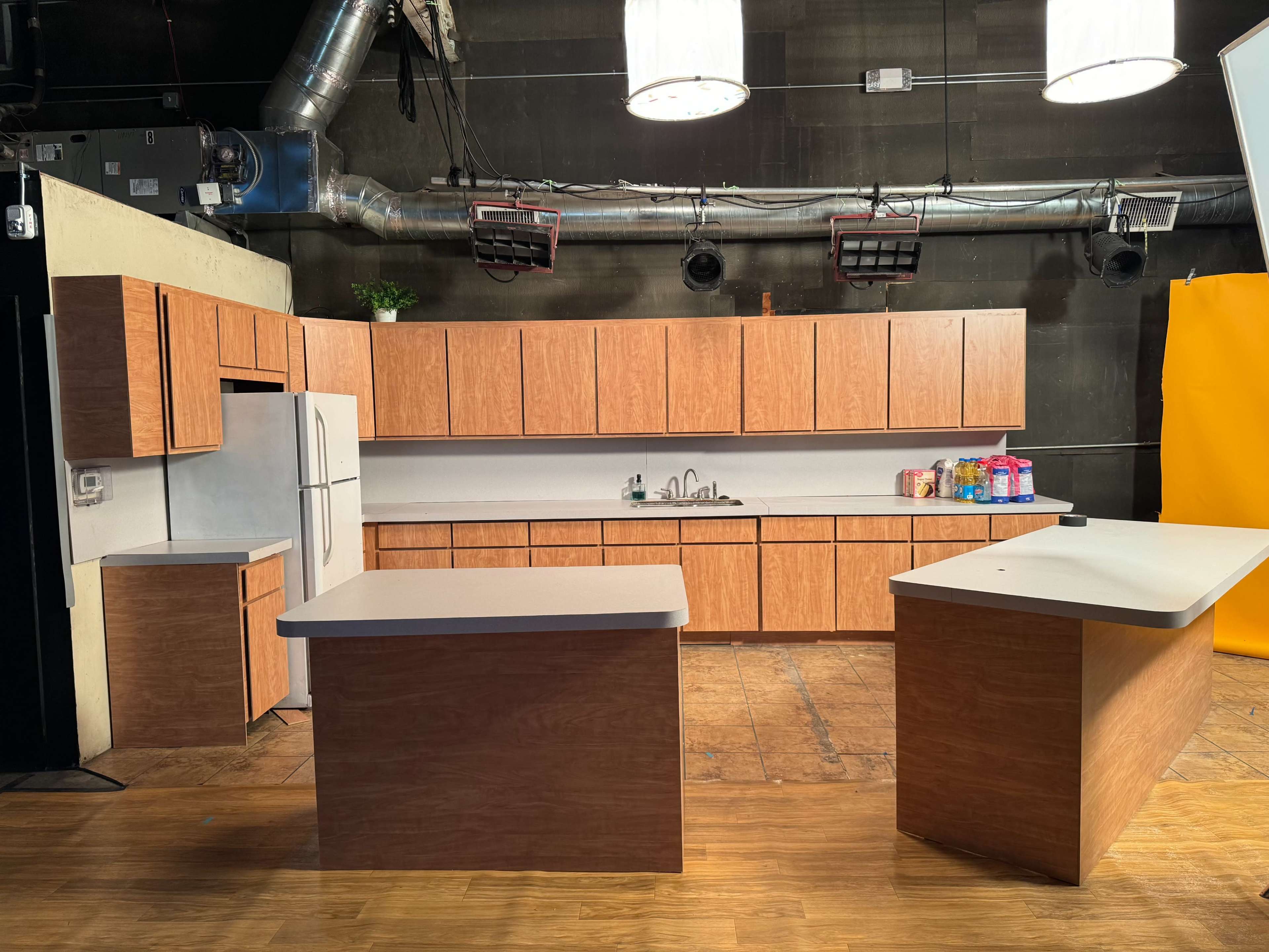 A kitchen set features wooden cabinetry, a refrigerator, and a central island under bright studio lights.