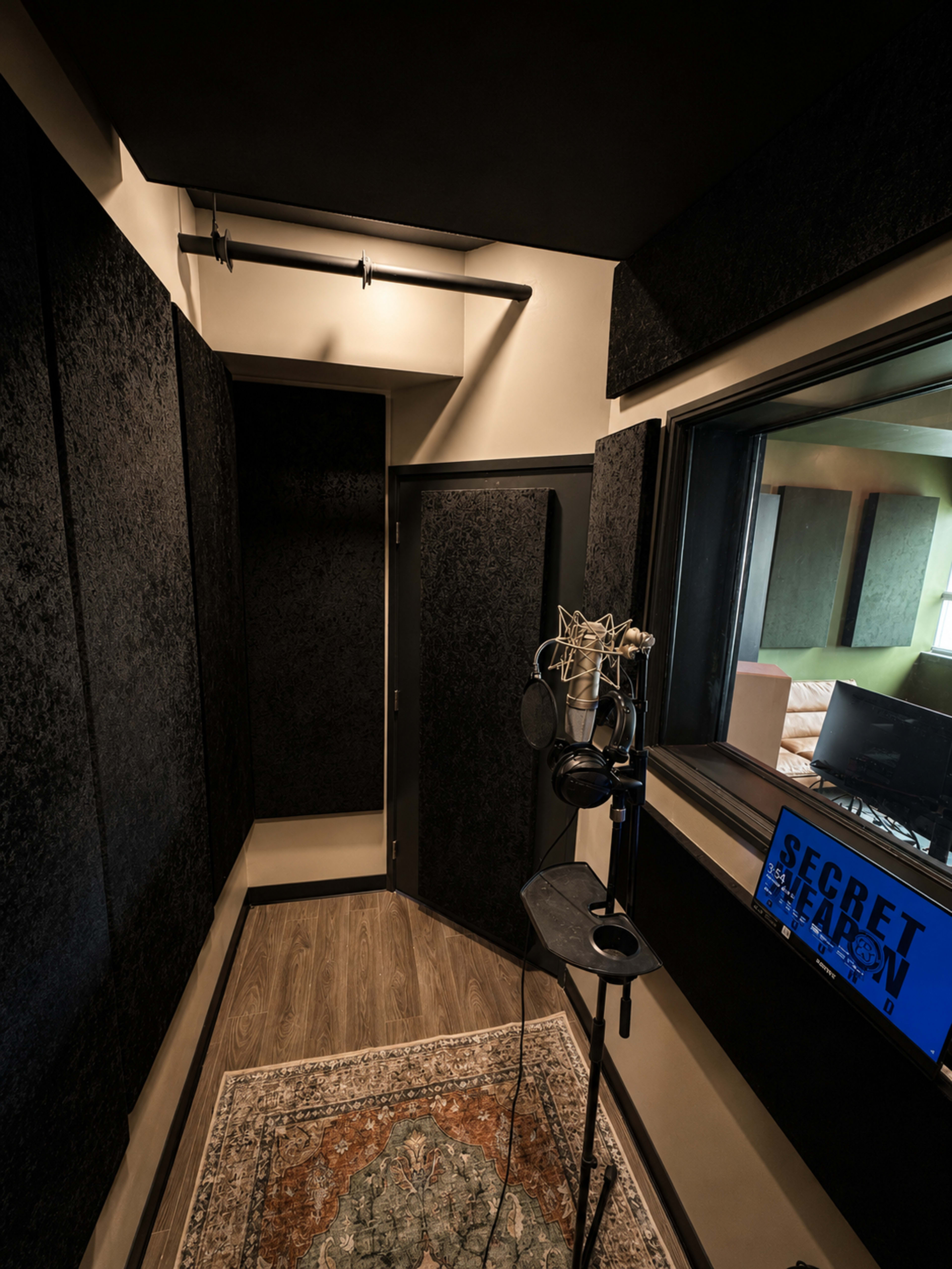 A soundproof recording studio features dark acoustic panels, a microphone on a stand, and a window to an adjacent room with soft seating.