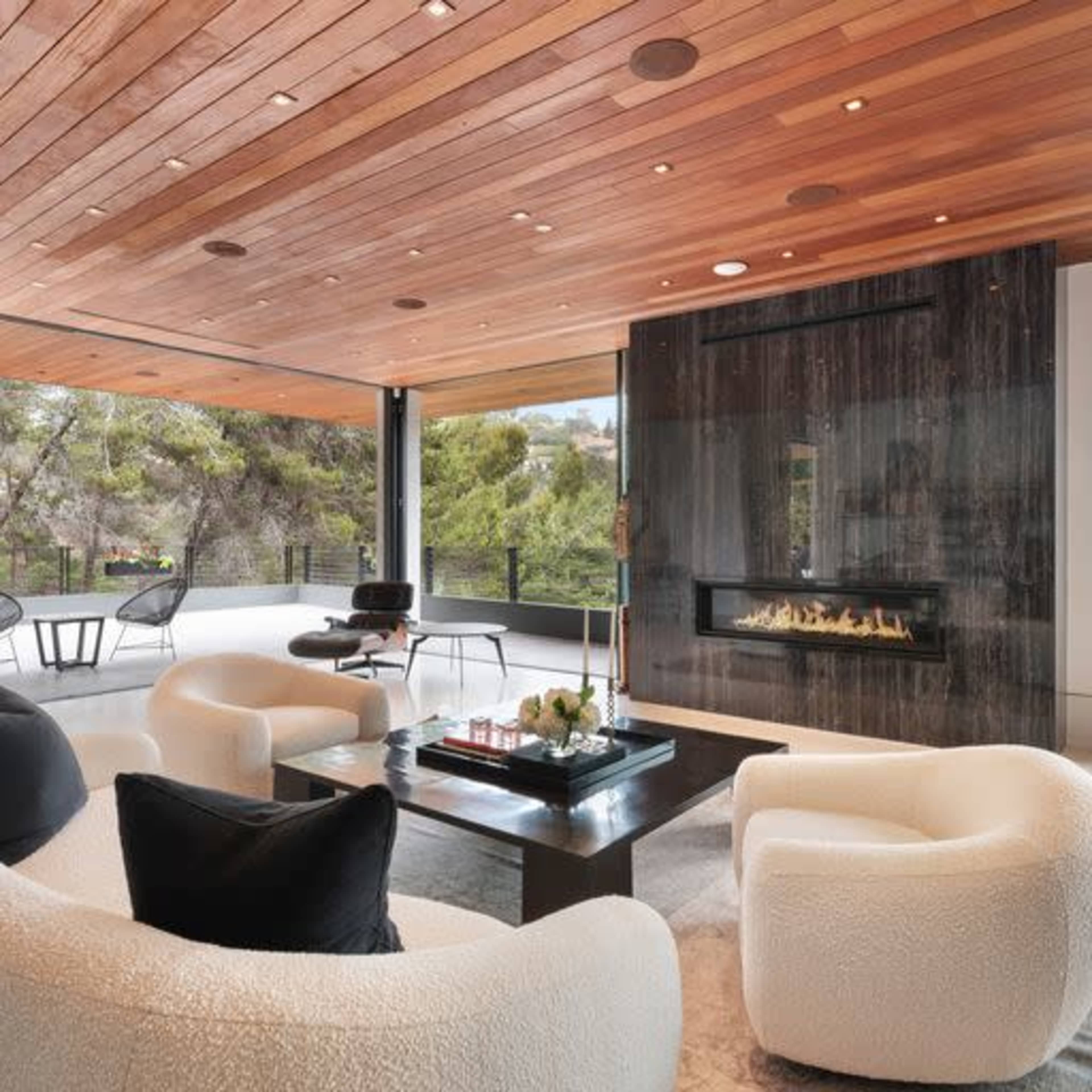 The scene features a modern living room with large windows, wooden beams on the ceiling, minimalist furniture, and a sleek fireplace.