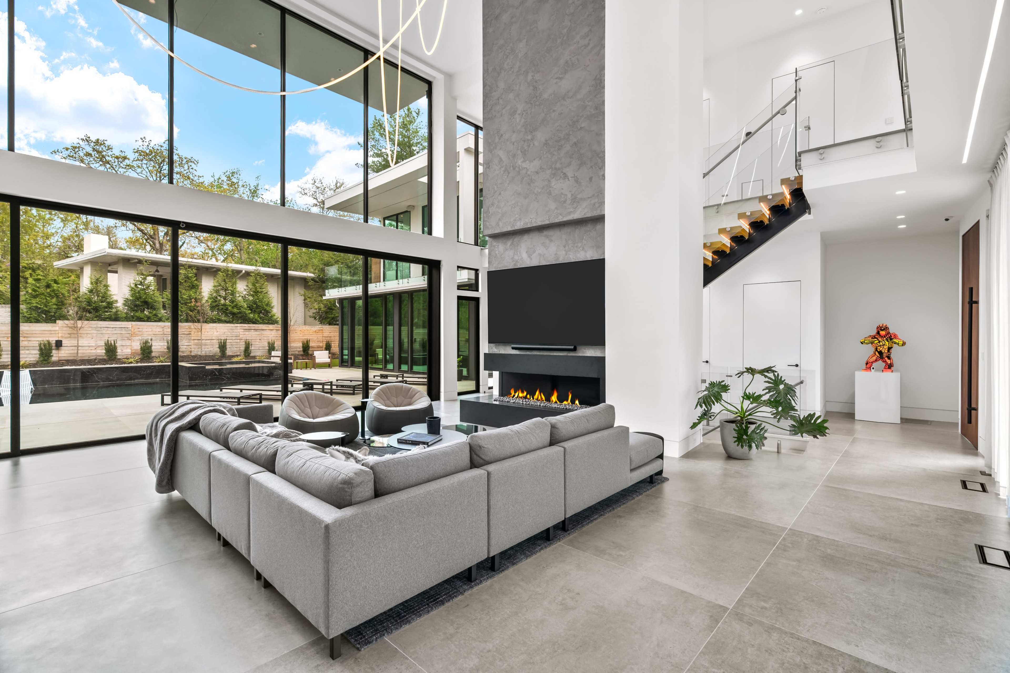 The image shows a modern living room with large glass windows, a gray sectional sofa, a sleek fireplace, and an open staircase leading to an upper level.
