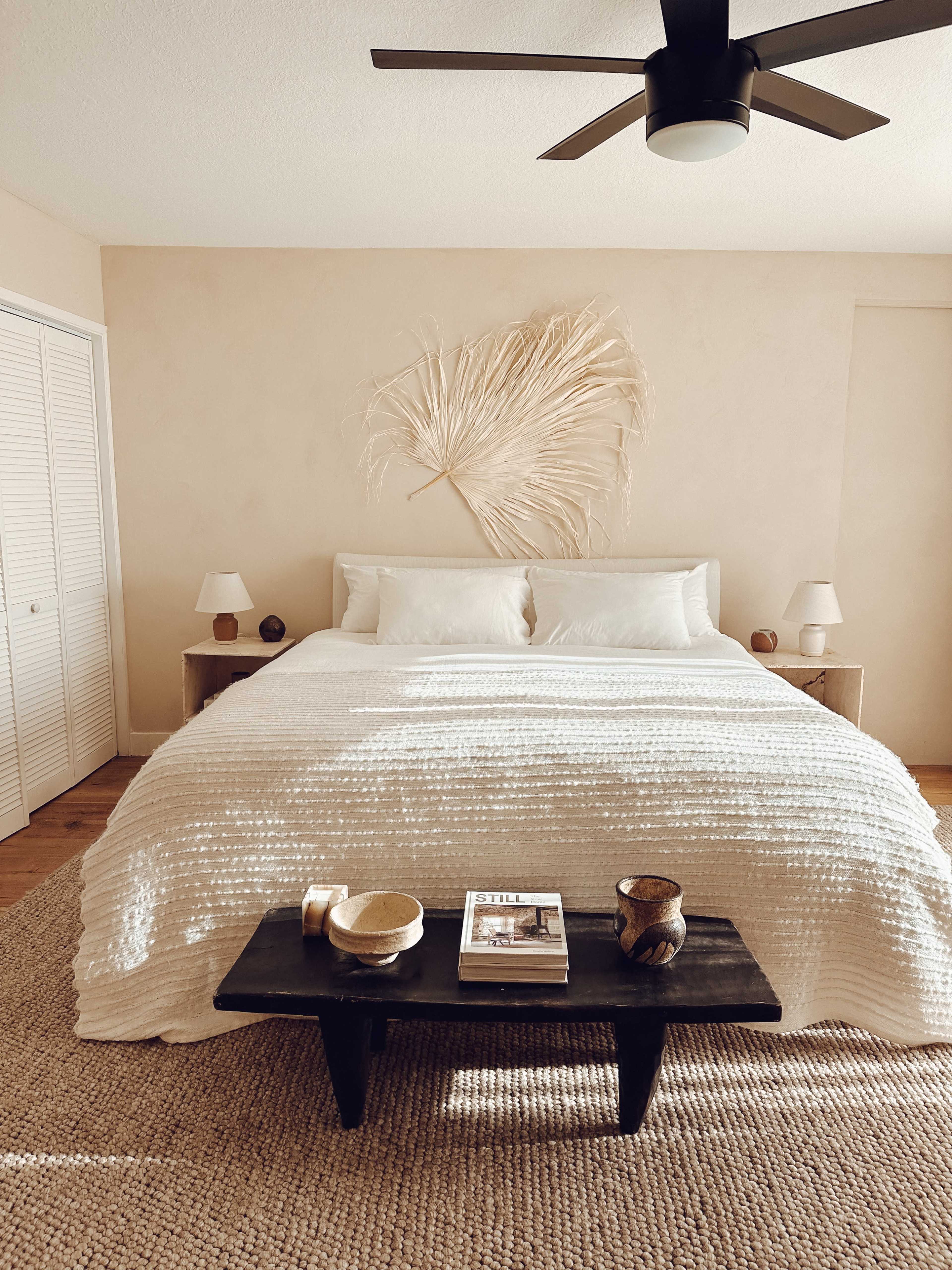 A serene bedroom with a large bed, decorative wall art of a dried palm leaf, and a small black coffee table in front.
