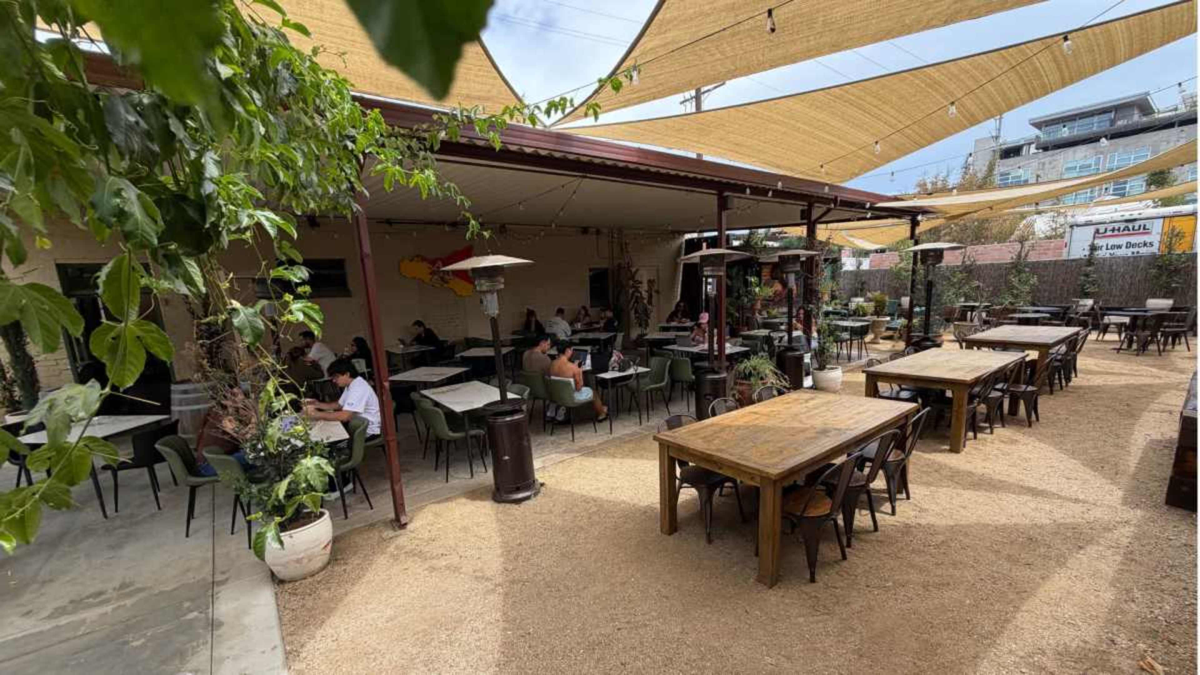 The image shows an outdoor dining area with several wooden tables, a shaded canopy overhead, and patrons seated among greenery and decor.