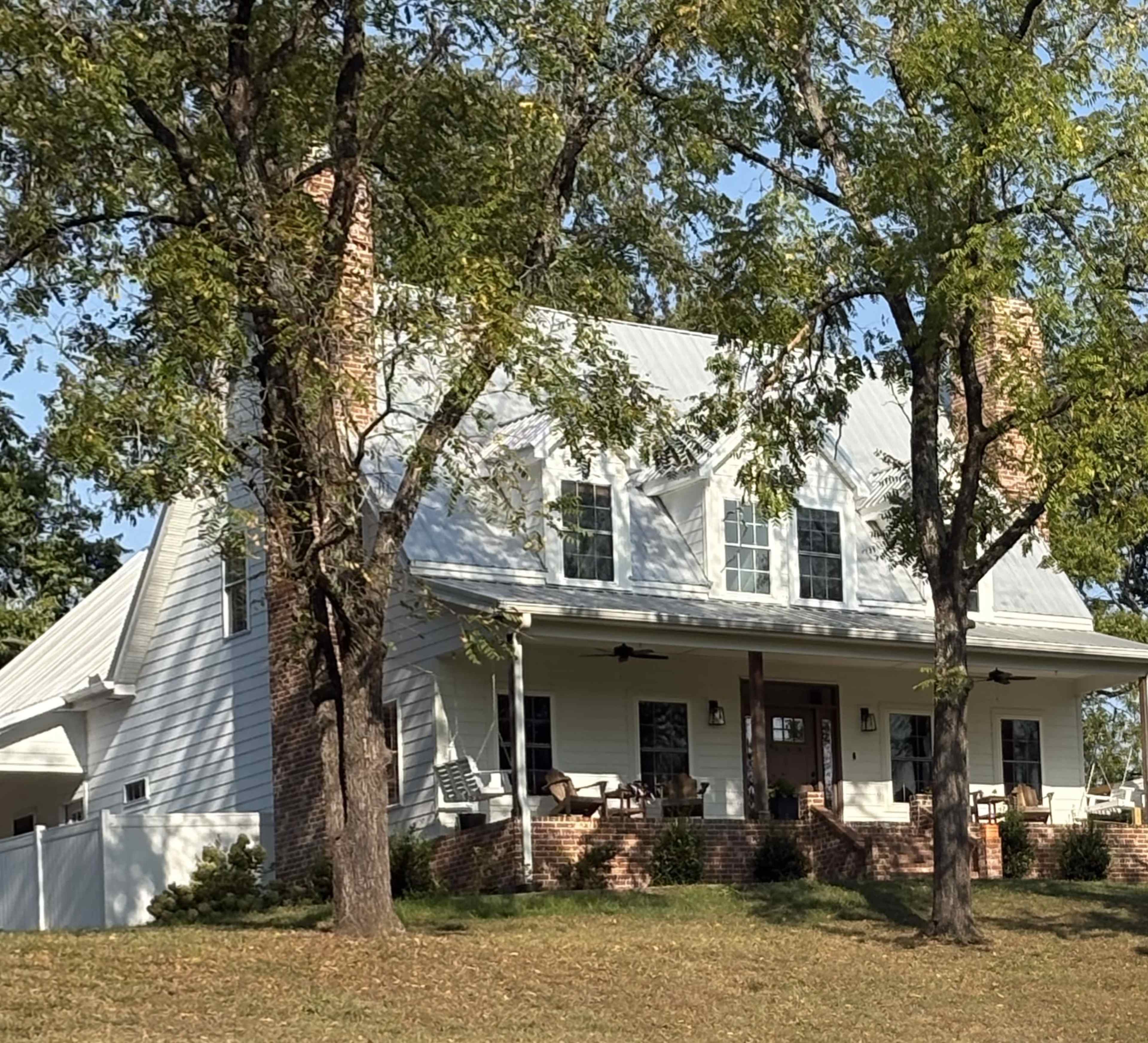 A two-story white house with a metal roof and brick foundation is surrounded by trees on a grassy slope.