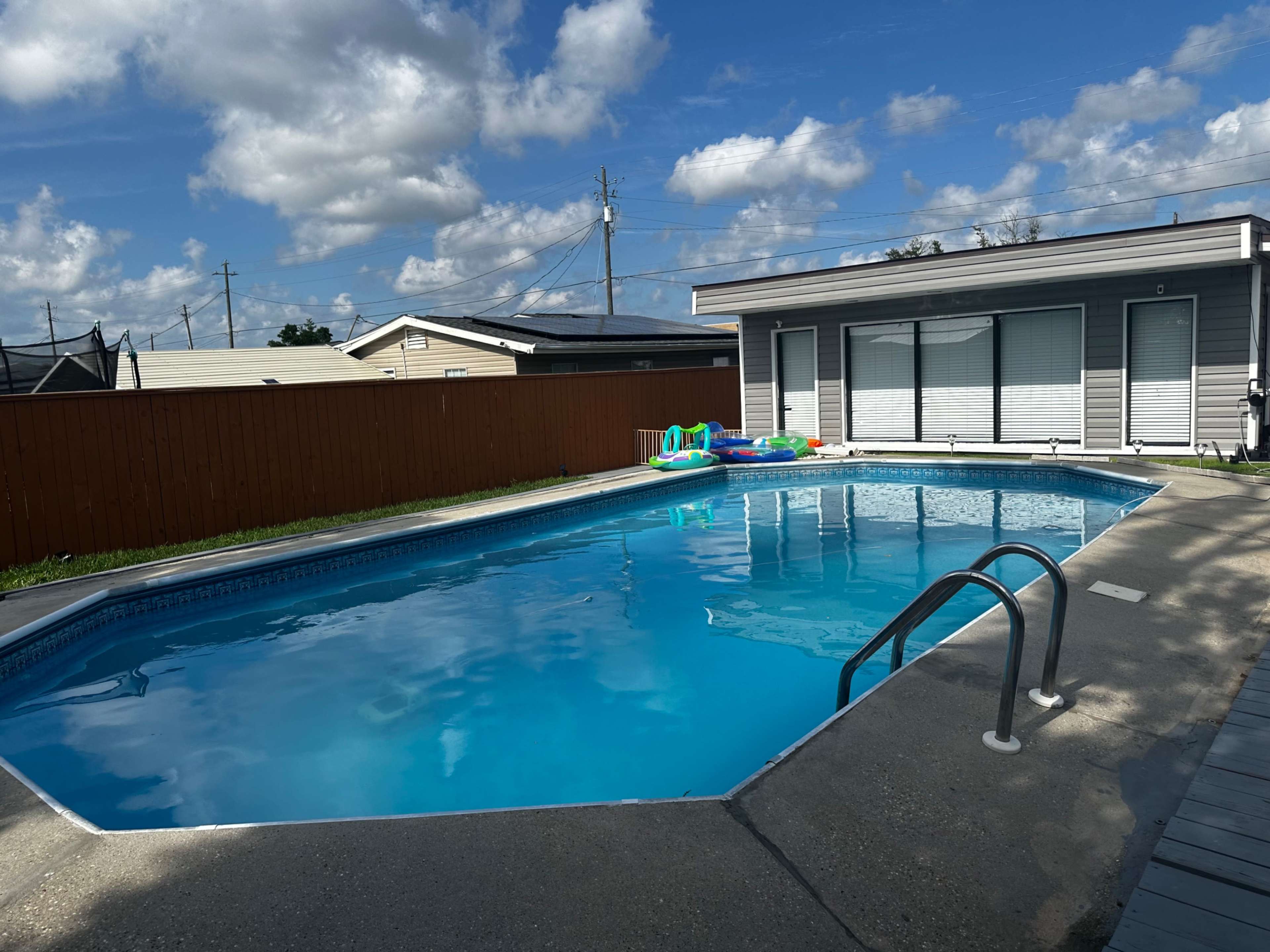 The image shows a rectangular swimming pool with clear blue water and a wooden fence in the background, next to a modern building with large windows.