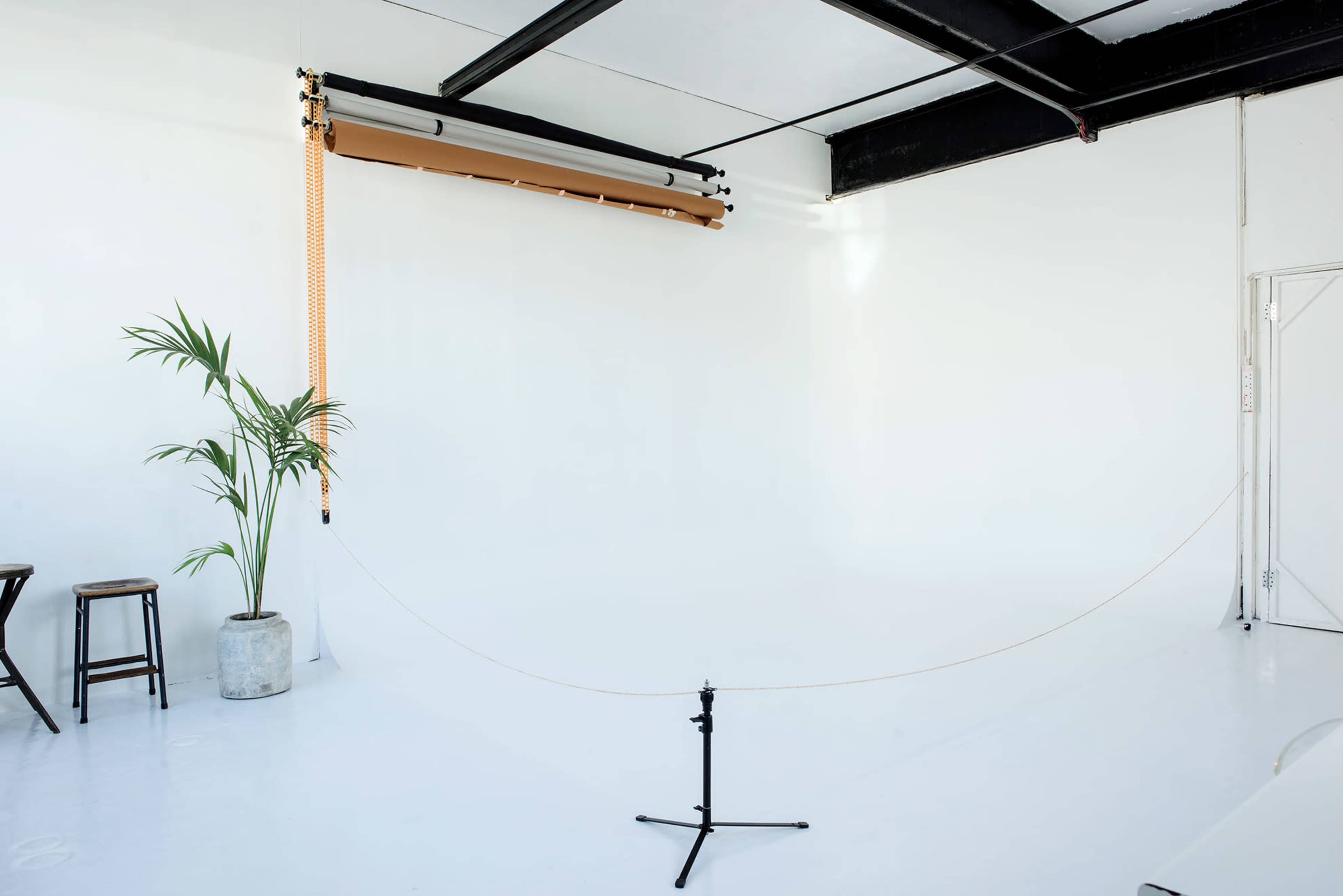 A minimalist studio space with a large roll of backdrop paper, a potted plant, and two wooden stools against a white wall.
