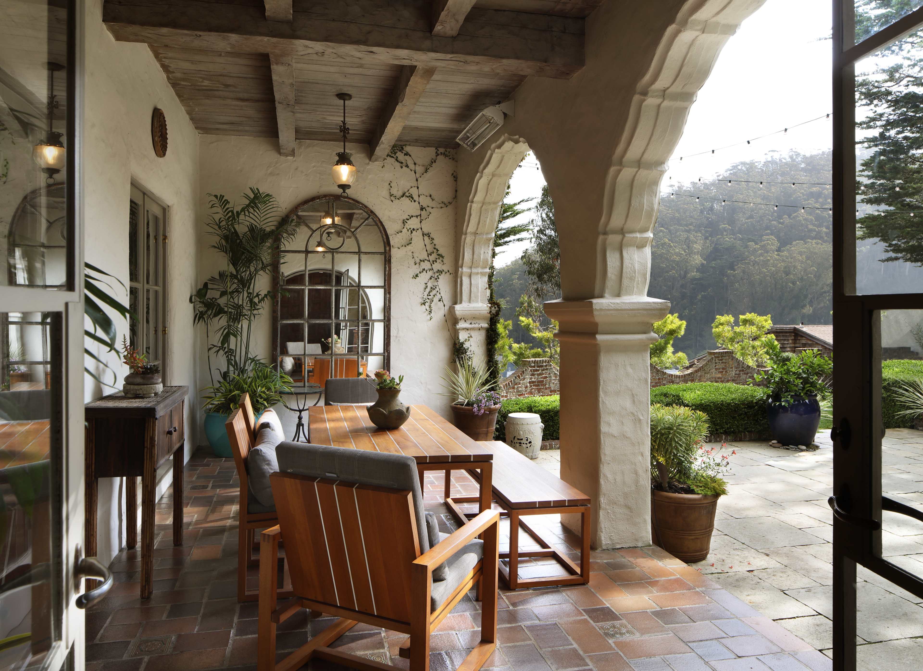A covered outdoor patio with wooden furniture and potted plants, framed by arched columns and large windows overlooking a garden.