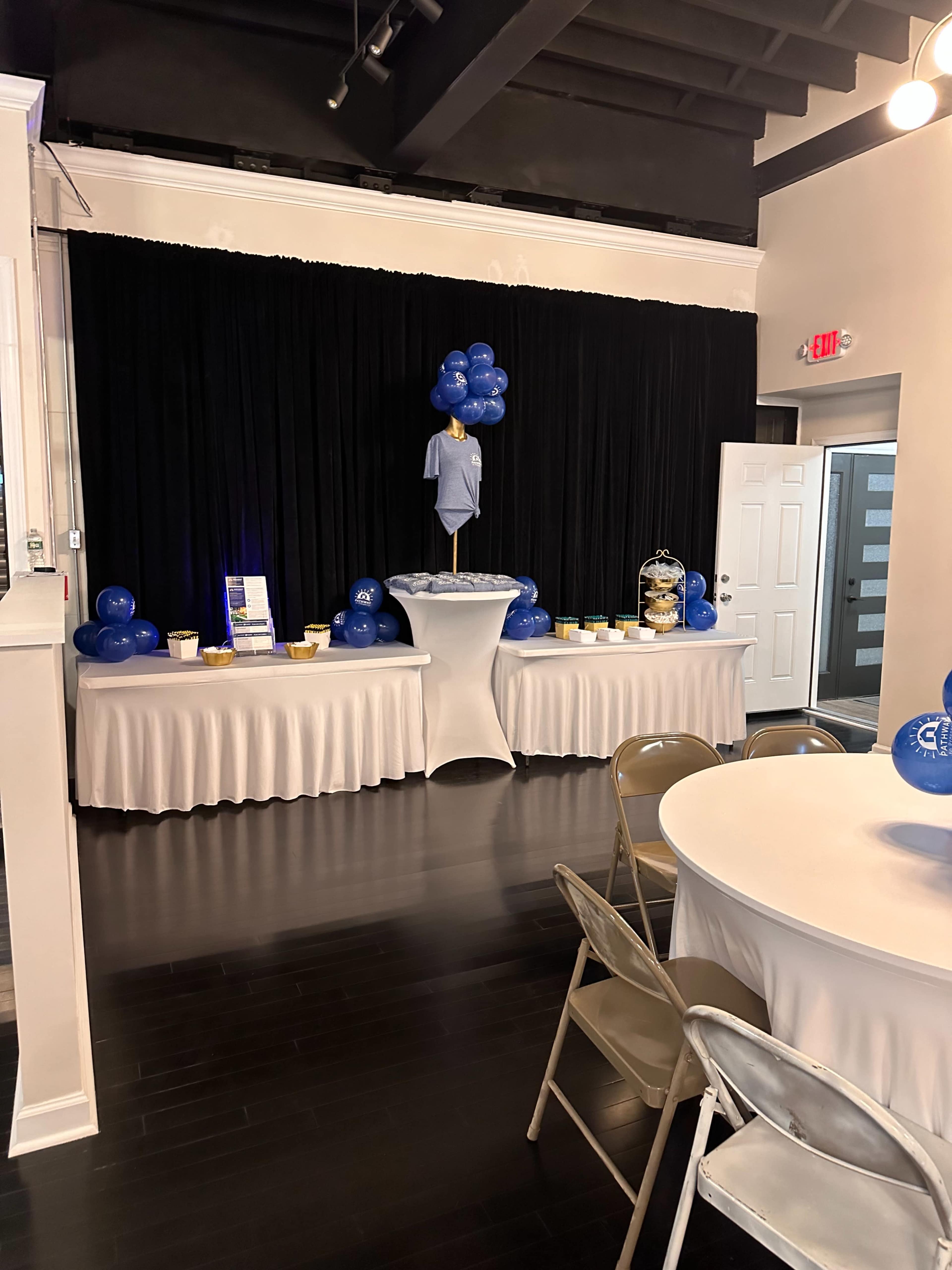 A table adorned with a blue balloon arrangement and a display shirt is set against a black backdrop in a well-lit event space.