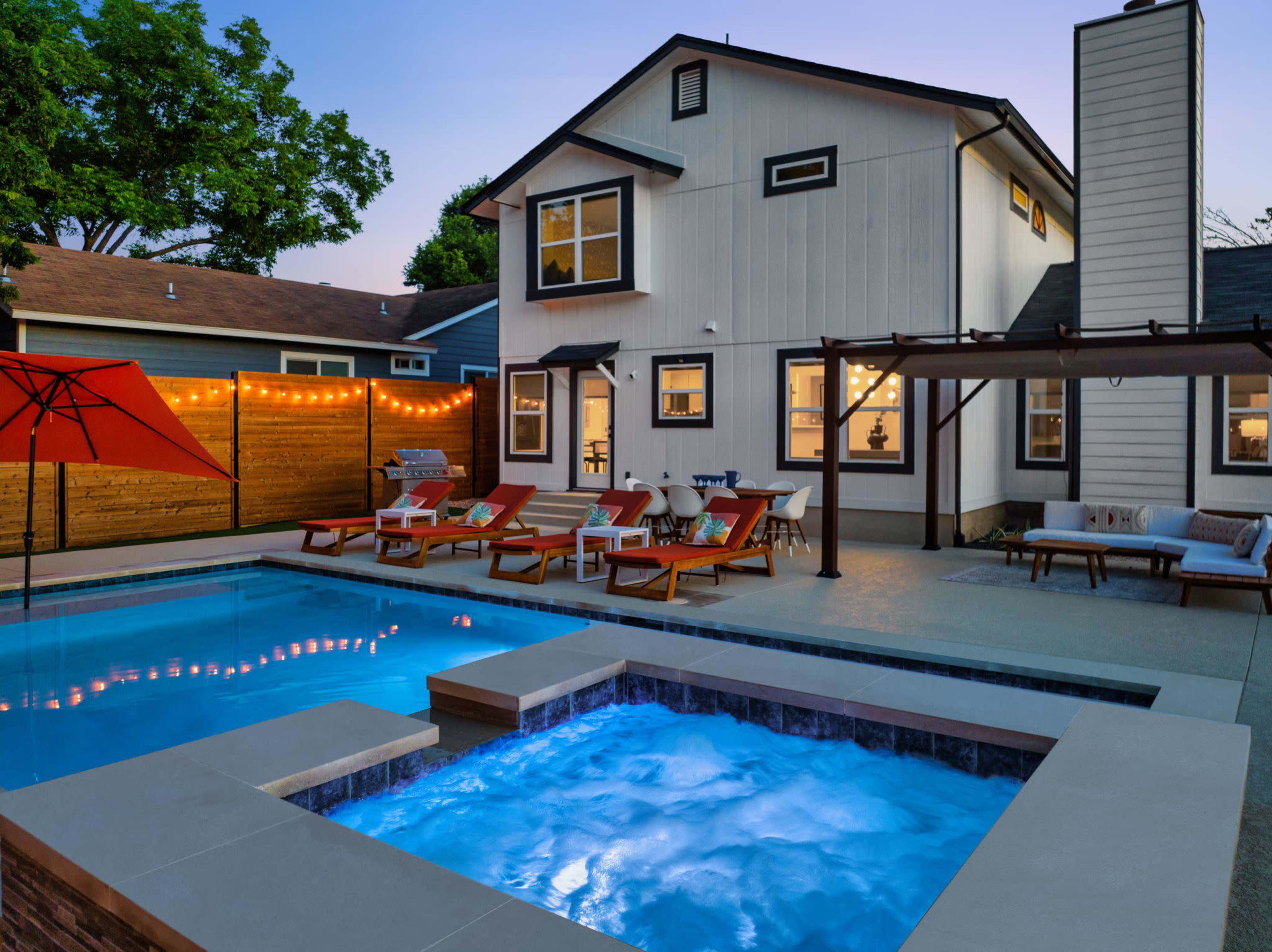 A modern house with a pool and spa features lounge chairs and an umbrella in the backyard, illuminated by string lights at dusk.