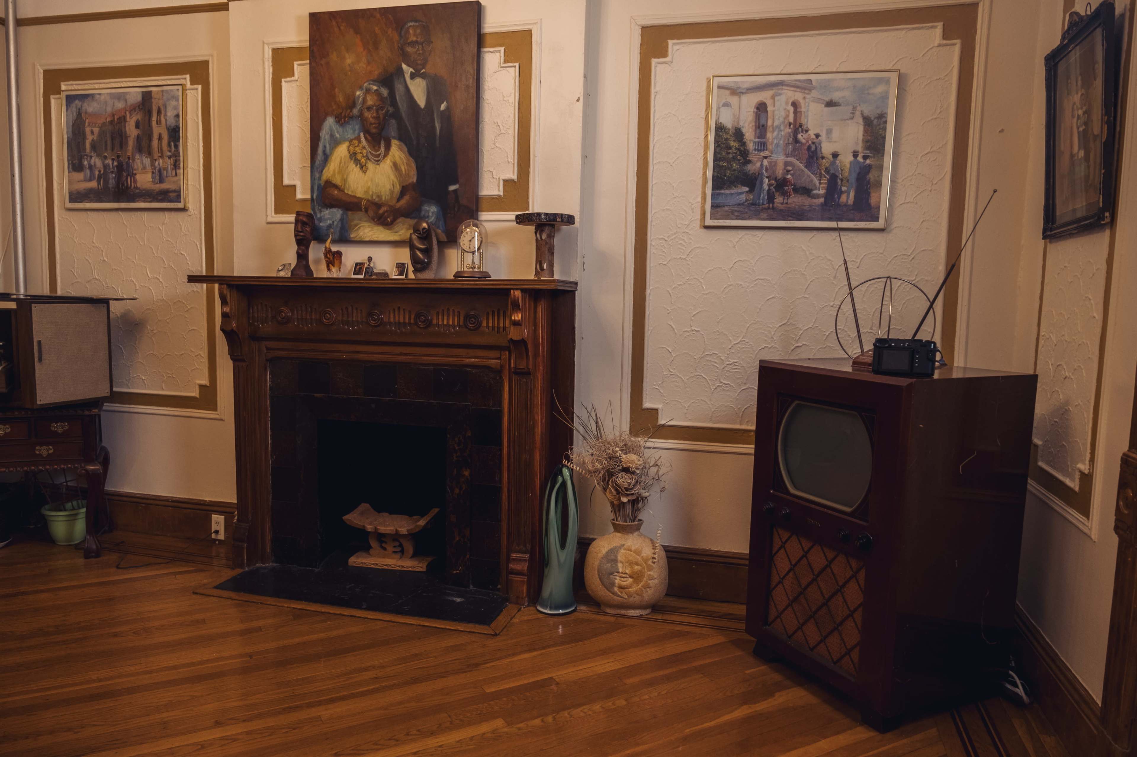 The image shows a living room with a wooden fireplace mantel, vintage artwork on the walls, and a classic television set on the right.