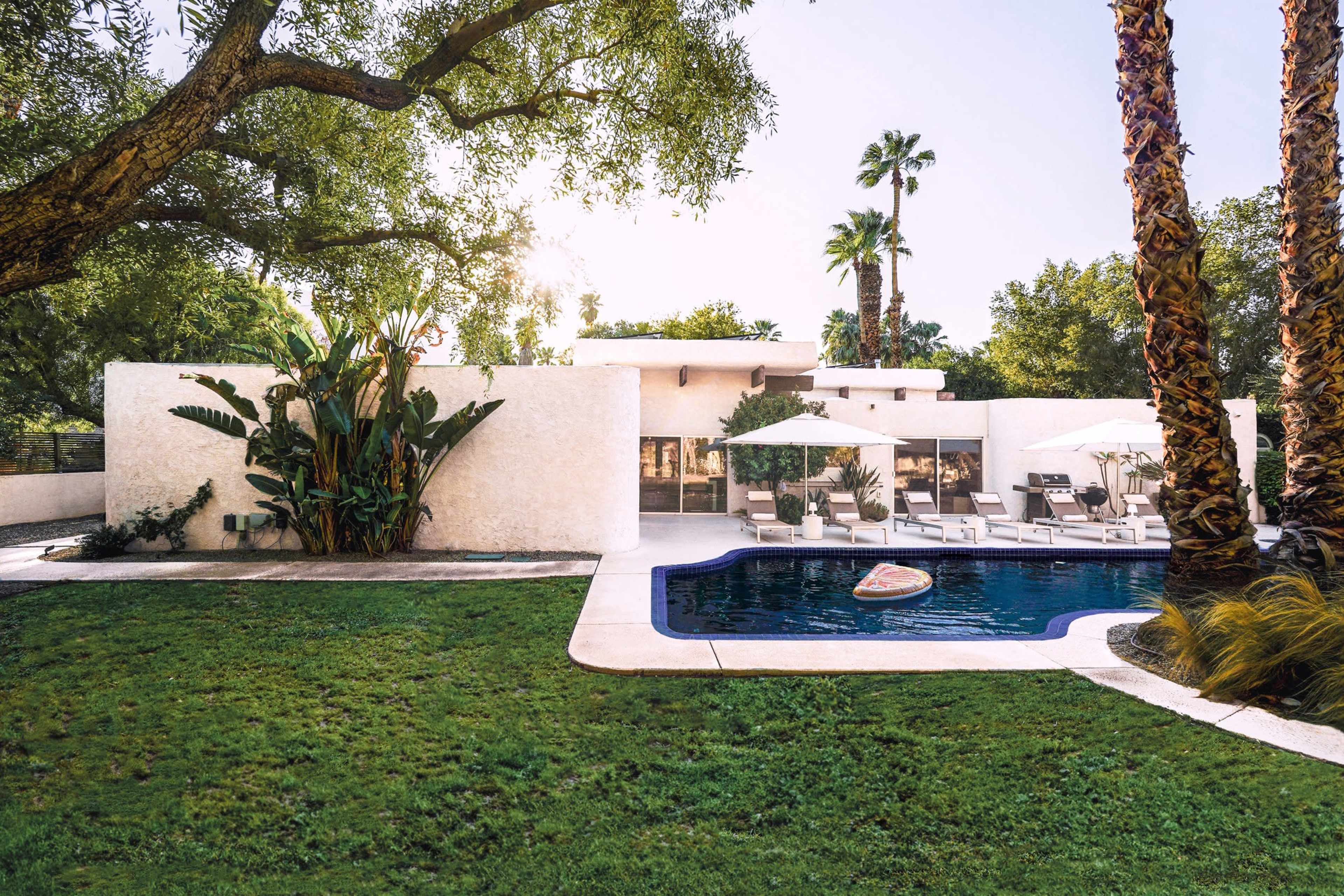 A modern house with large windows is surrounded by palm trees and features a swimming pool with lounge chairs and umbrellas in the backyard.