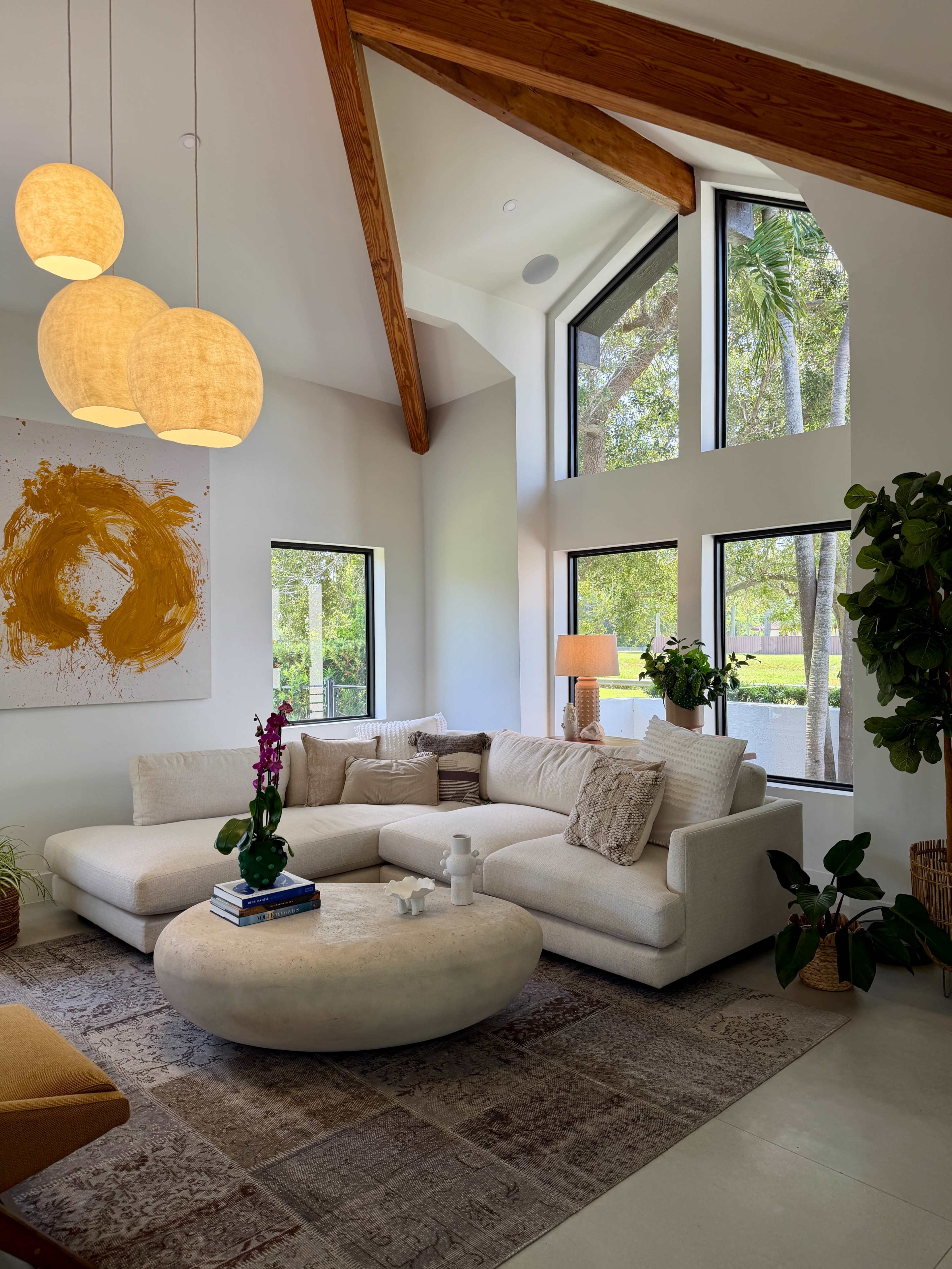 The image shows a modern living room with a sectional sofa, round coffee table, large windows letting in natural light, and decorative plants.