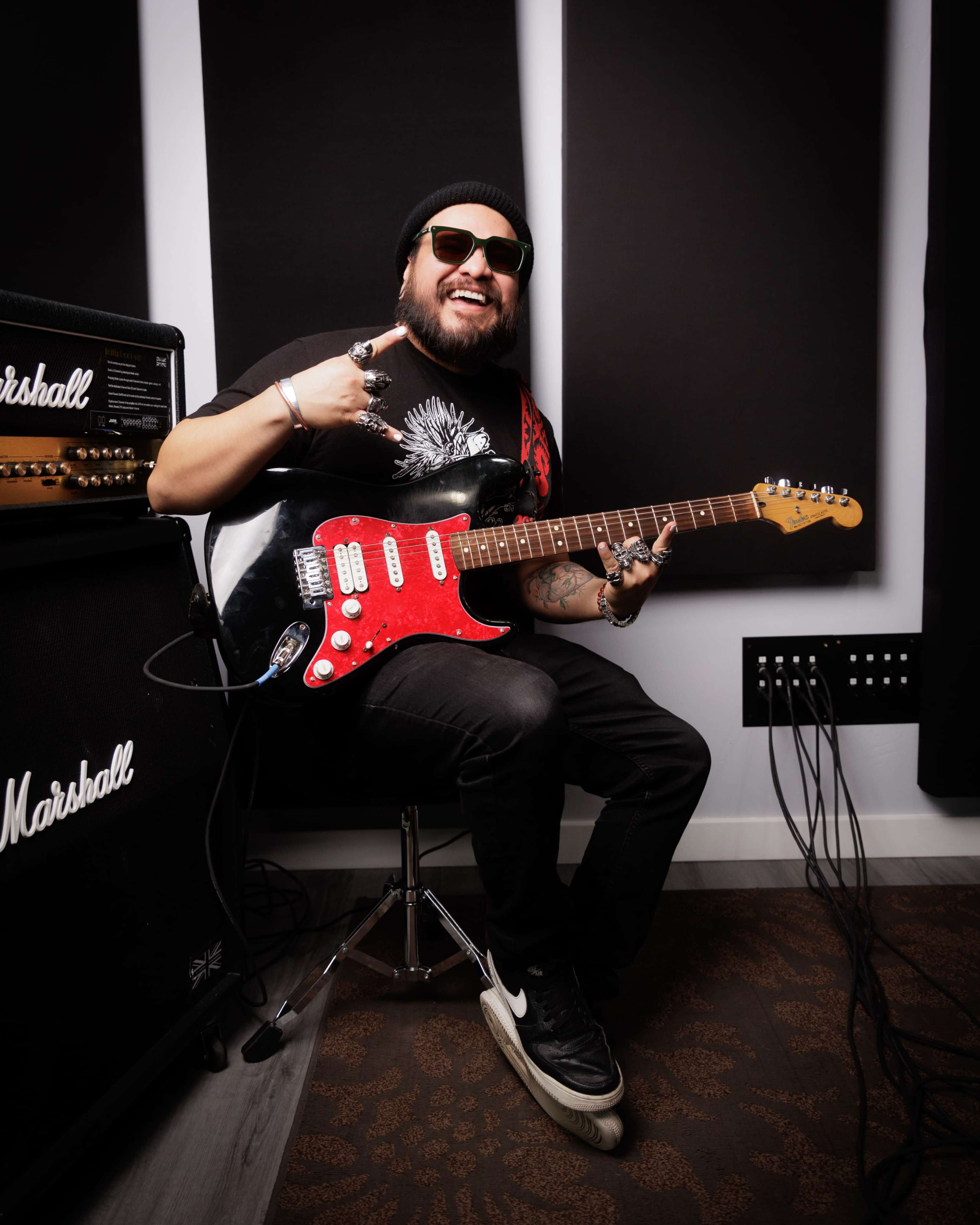 A musician plays a red electric guitar while seated next to a Marshall amplifier in a recording studio.